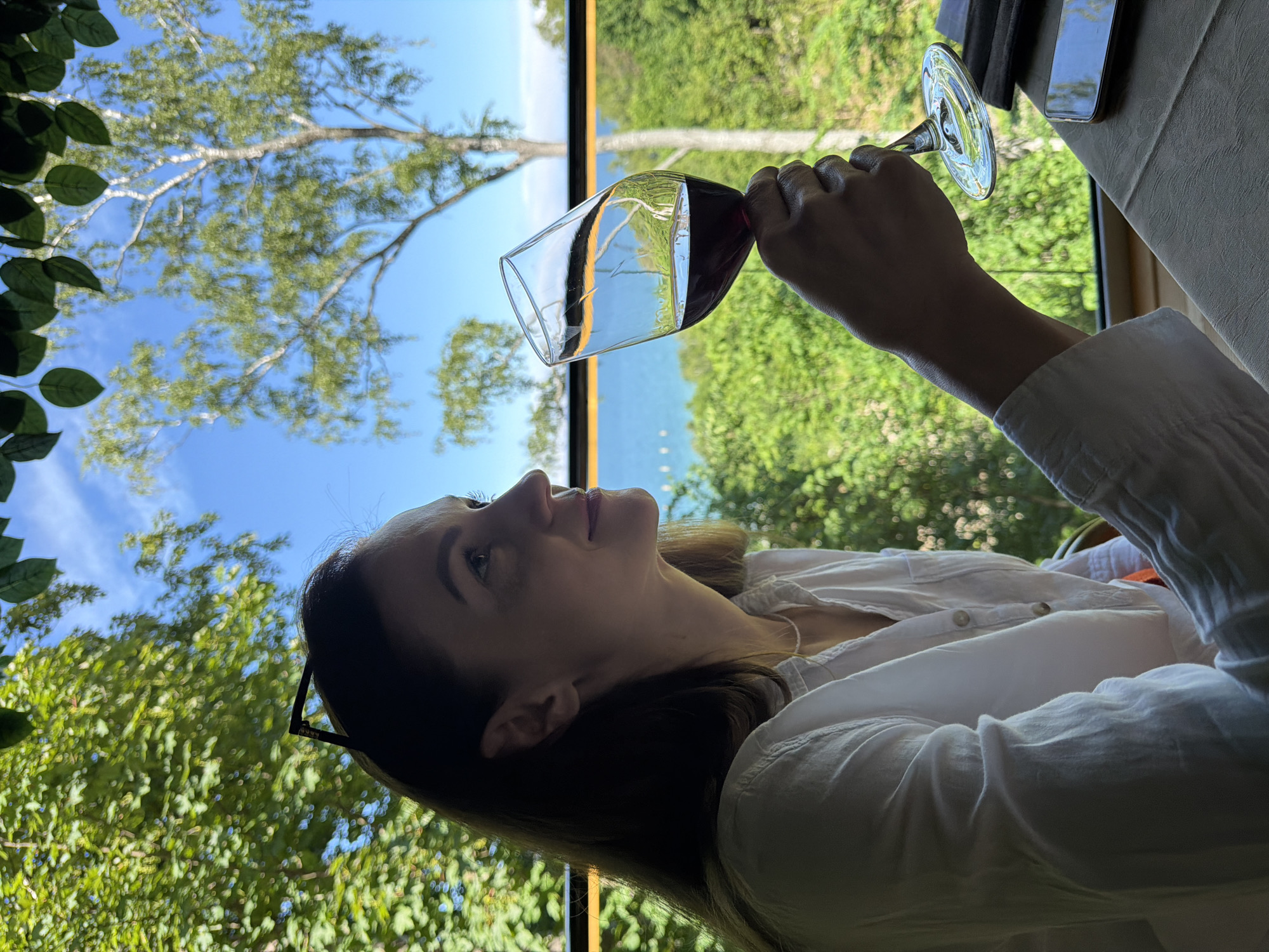 A woman in a white shirt is holding a glass of red wine in her right hand, looking out at a scenic view. She is seated at a table, with lush green trees and a bright blue sky visible through a large window. The scene appears to be in a restaurant or outdoor seating area, possibly overlooking a body of water. The woman has sunglasses perched on her head and appears to be relaxed and enjoying the moment. The table is set with a tablecloth and a smartphone is visible. The overall atmosphere is one of tranquility and enjoyment, enhanced by the natural beauty of the surroundings. It is likely the image was taken in Svetlogorsk, Russia, given the presence of birch trees and the proximity to the Baltic Sea, a common sight in the resort town.