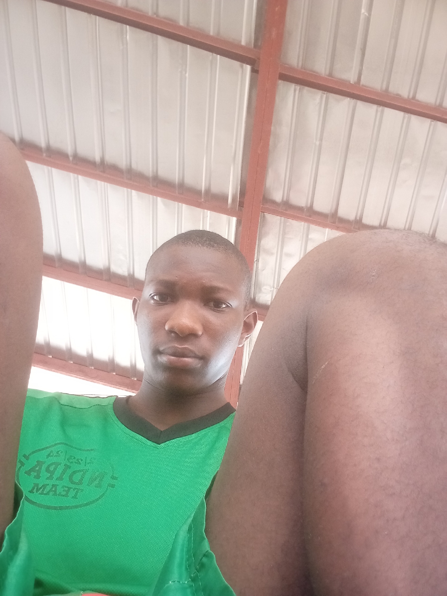 A dark-skinned male is in the foreground, wearing a green t-shirt. A body part (likely a thigh or shoulder) is adjacent to the subject’s right side. The background shows a metal or corrugated iron ceiling with red-painted framing. The location is unconfirmed as Onitsha, Nigeria.