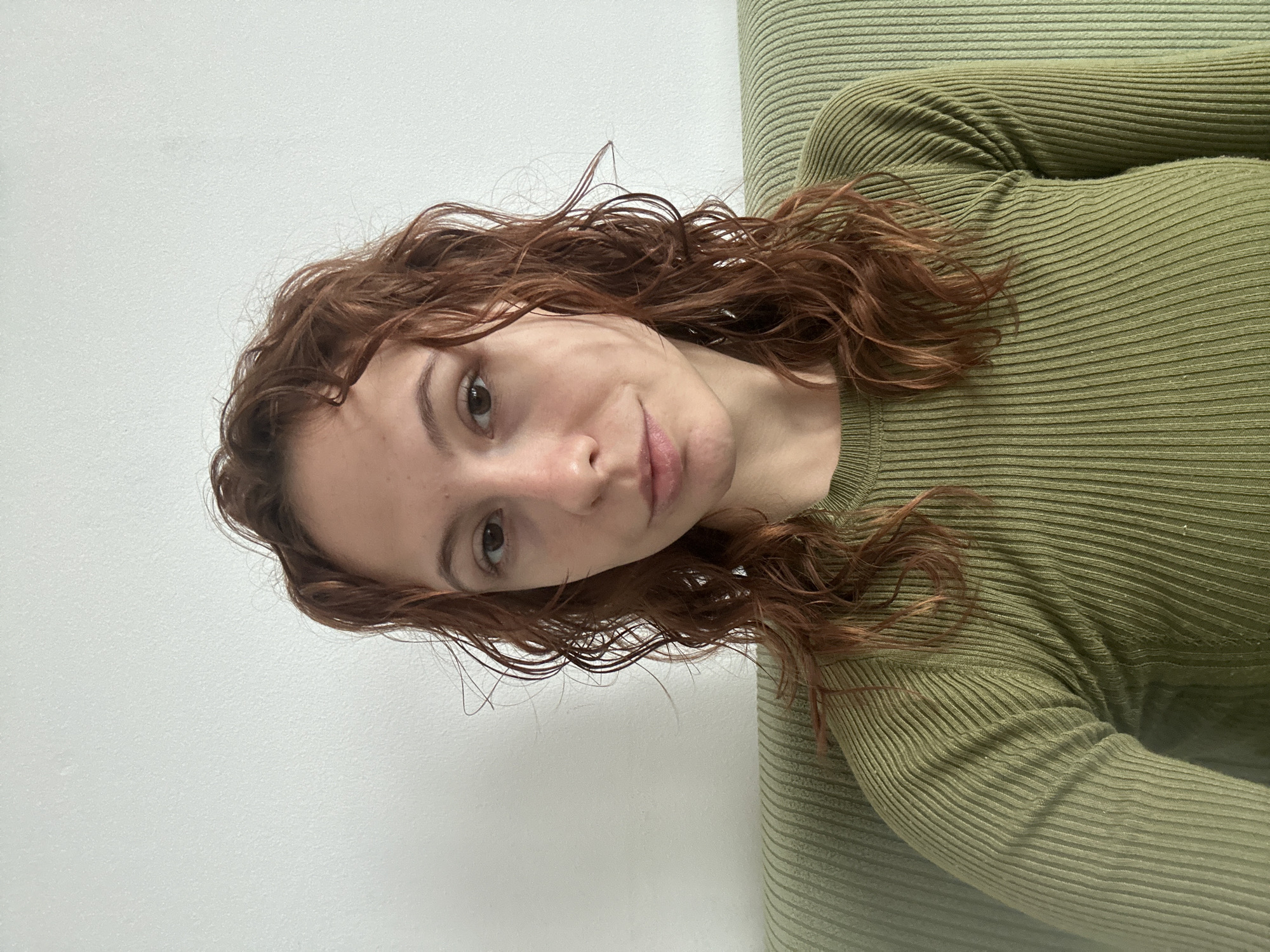 A young woman with light skin and reddish-brown curly hair is centered in the image. She is wearing a ribbed, olive-green long-sleeved top. Her head is tilted to the right, and she is looking directly at the camera. The background is divided between a light-colored wall on the left and a textured, presumably upholstered, surface in a similar shade of green to her shirt on the right. No specific elements indicate that this photo was taken in Novi Sad, Serbia, or provide any other geographical information.