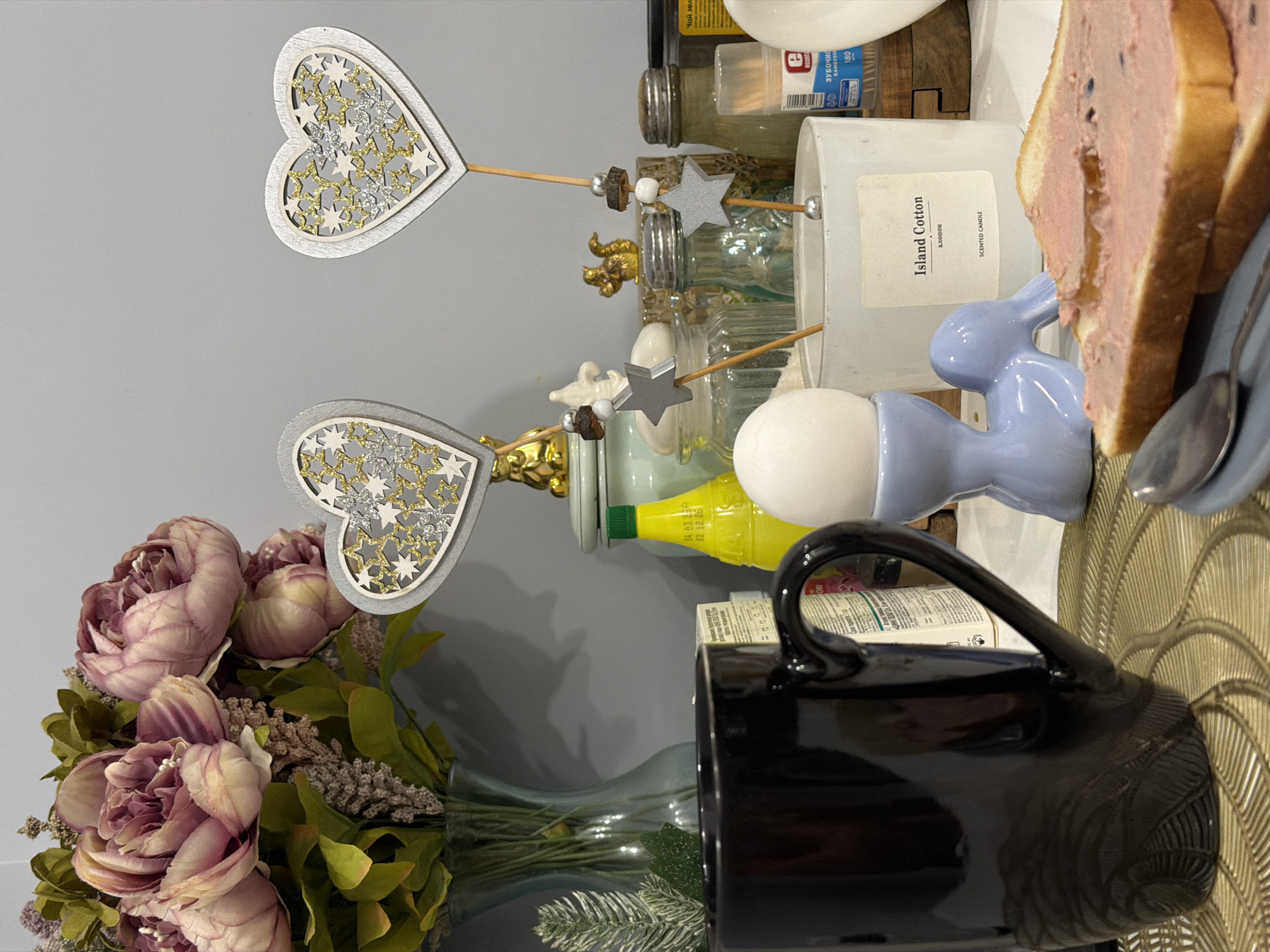 This image presents a still life arrangement with several objects on a surface. In the foreground, there's a bouquet of pink and purple flowers in a vase, beside a black mug with a visible handle. Behind these, there are two heart-shaped decorations on sticks, placed above glass jars, a candle labeled "Island Cotton", a blue ceramic bunny figure next to a white egg on an egg holder, and a slice of bread. A spoon lies on a plate. The overall environment suggests an indoor setting.