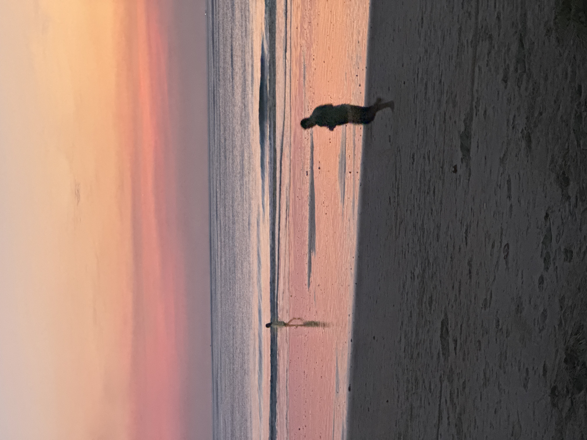 A photograph taken at sunset on the beach in Kuta Selatan, Indonesia, shows two figures in silhouette against a gradient sky of orange, pink, and purple hues. The ocean, reflecting the sky's colors, stretches to the horizon. On the wet sand near the water's edge, a person walks away from the camera. Further inland on the darker sand, another person is captured in mid-stride, appearing to be running. The foreground of the image is dominated by the textured, dark sand of the beach.