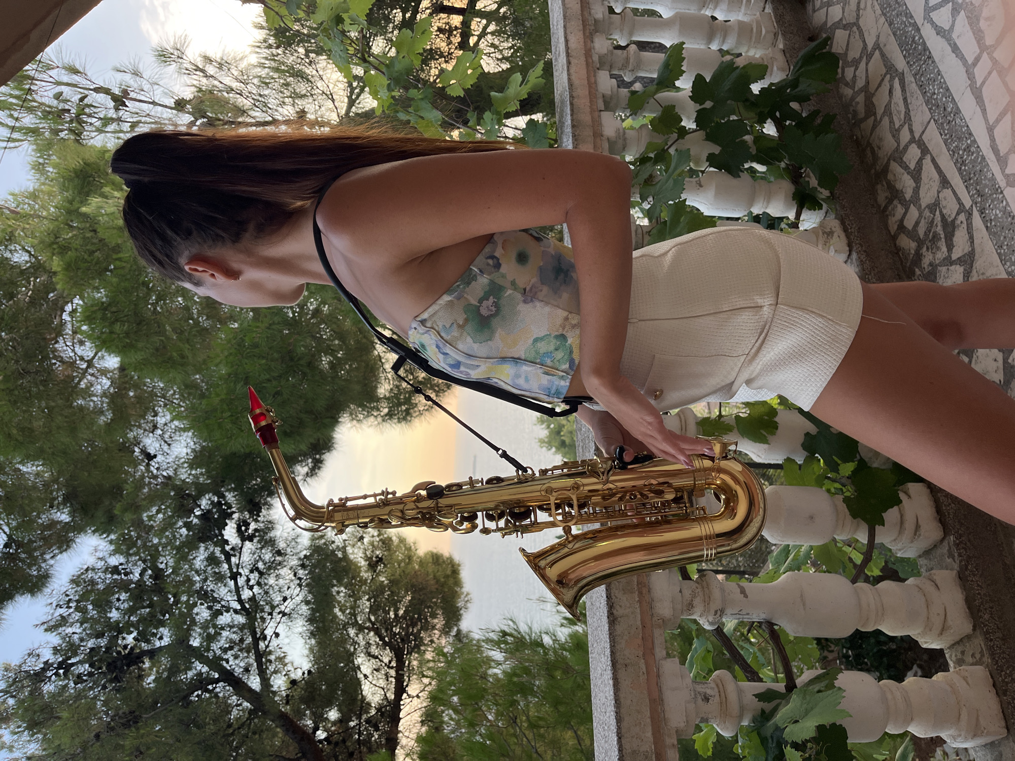 A woman stands on a balcony in Šušanj, Montenegro, holding a saxophone. The balcony has a white railing and a tiled floor. In the background, trees and the sea are visible, with a ship on the horizon. The sky is clear with a hint of sunset. The woman is wearing a floral-patterned strapless top and white shorts. Her hair is pulled back in a ponytail. She is looking out towards the sea, and her hands are positioned on the saxophone as if she is about to play. The overall scene suggests a serene and picturesque moment.