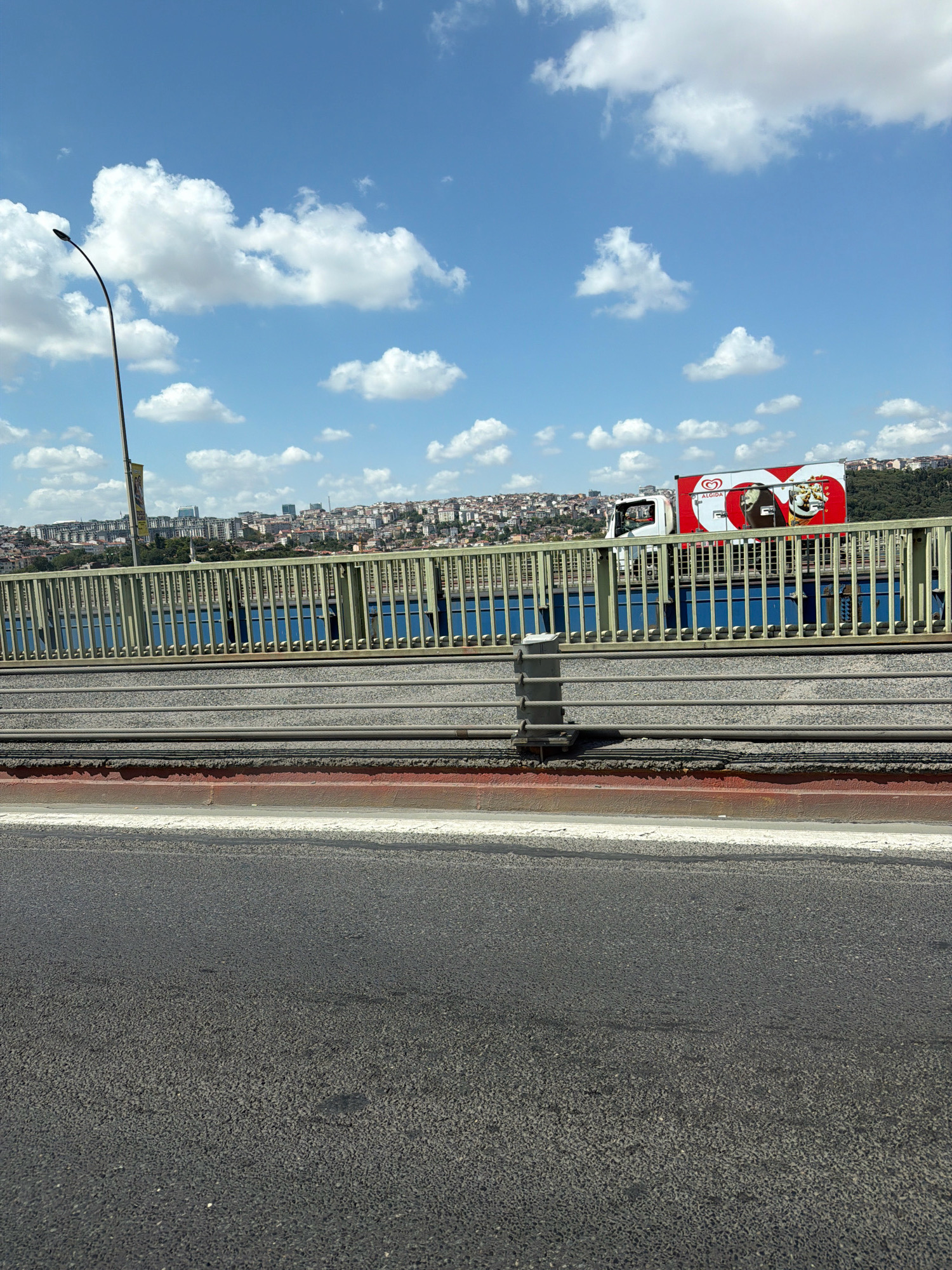 A white truck with an "Algida" advertisement, featuring ice cream images and a heart logo, is driving on a bridge over a body of water. The truck has a red and white trailer. In the background, a cityscape with numerous buildings and a hill covered in trees is visible. The sky is blue with scattered white clouds. A street lamp is present on the left side of the image. The foreground shows a road and a metal railing. This scene is likely in Eyüpsultan, Turkey, given the presence of the bridge and cityscape which resembles the Bosphorus strait.
