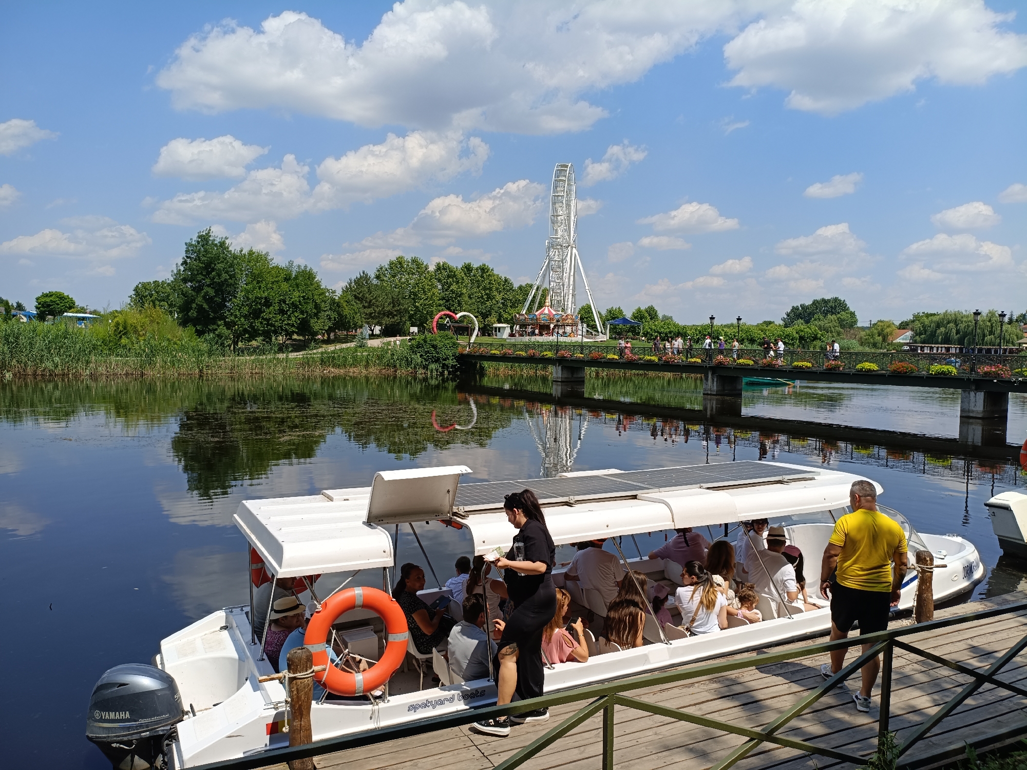 The image depicts a sunny day at a waterfront attraction in Comana, Romania. A white passenger boat, branded "Spakyard Boats" and powered by a Yamaha outboard motor, is docked at a wooden pier. The boat is filled with approximately ten people, appearing to be tourists, enjoying a ride or waiting to board. Some passengers are seated under a canopy, while others are exposed to the sun. An orange life preserver is visible on the side of the boat.
In the background, a pedestrian bridge spans across a calm body of water, adorned with colorful flowers. Beyond the bridge, a Ferris wheel and a carousel are prominent attractions, suggesting a recreational park or amusement area. The scene is framed by lush green trees and reeds along the water's edge, with a clear blue sky filled with fluffy white clouds overhead. The overall atmosphere is peaceful and recreational.