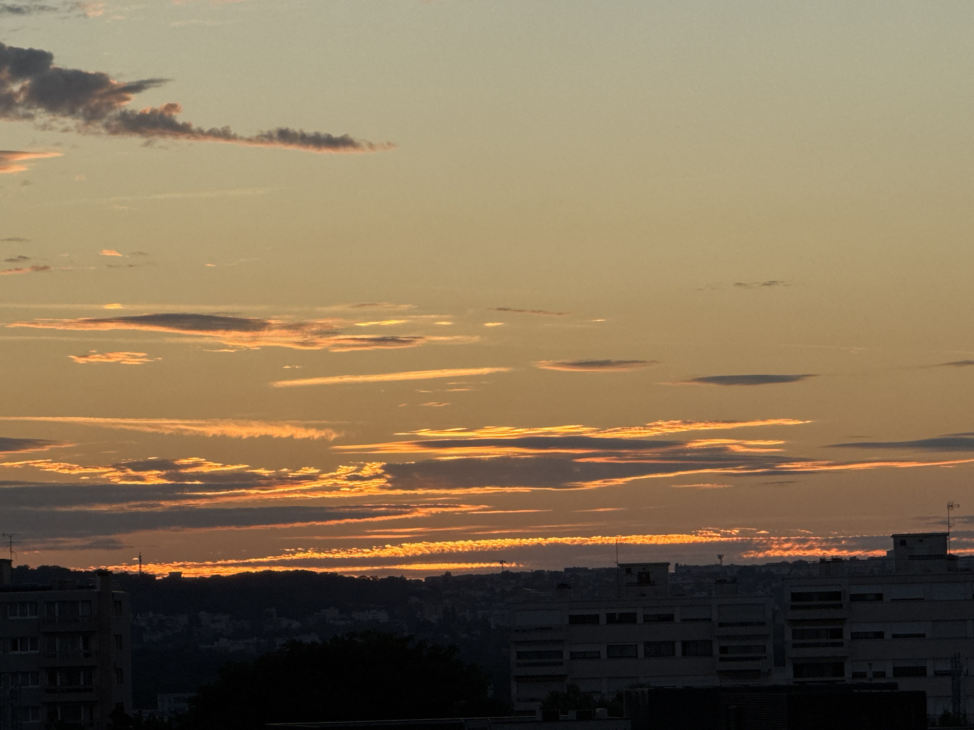 A cityscape is captured during sunset, with the sky ablaze with hues of orange and yellow. Long, thin clouds are scattered across the sky, illuminated by the setting sun, creating a dramatic visual effect. In the foreground, the silhouettes of buildings are visible against the vibrant sky. The buildings appear to be of varying heights and architectural styles, with some featuring multiple stories and windows. A dark silhouette of trees and a hill is present in the middle ground, obscuring the lower parts of the city. The overall scene evokes a sense of urban tranquility as day transitions into night.