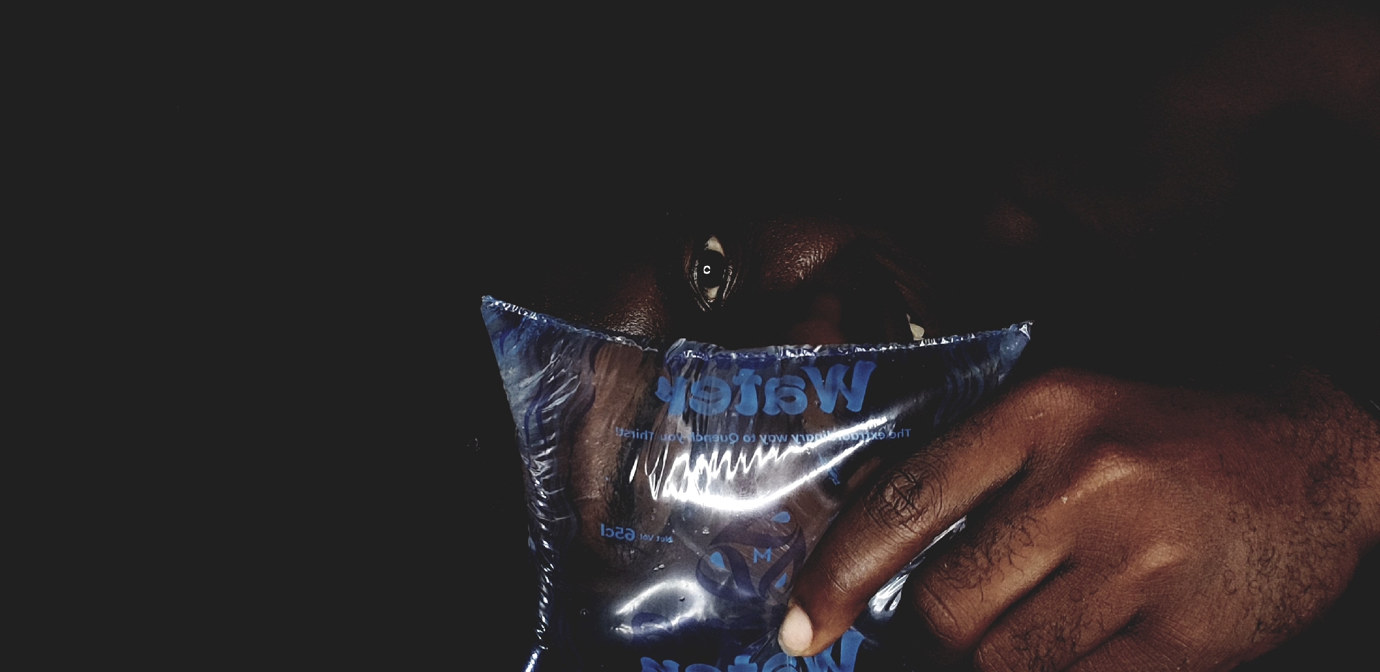 A close-up photograph depicts a person with dark skin holding a blue plastic water pouch. The pouch obscures the person's mouth and chin, with only one eye visible. The hand holding the pouch is also dark-skinned. The background is a dark, blurred tone, which could indicate a dimly lit indoor location. The words "Water" are printed in blue on the plastic bag.
There is nothing in the photograph that indicates the city of Narayi, Nigeria.