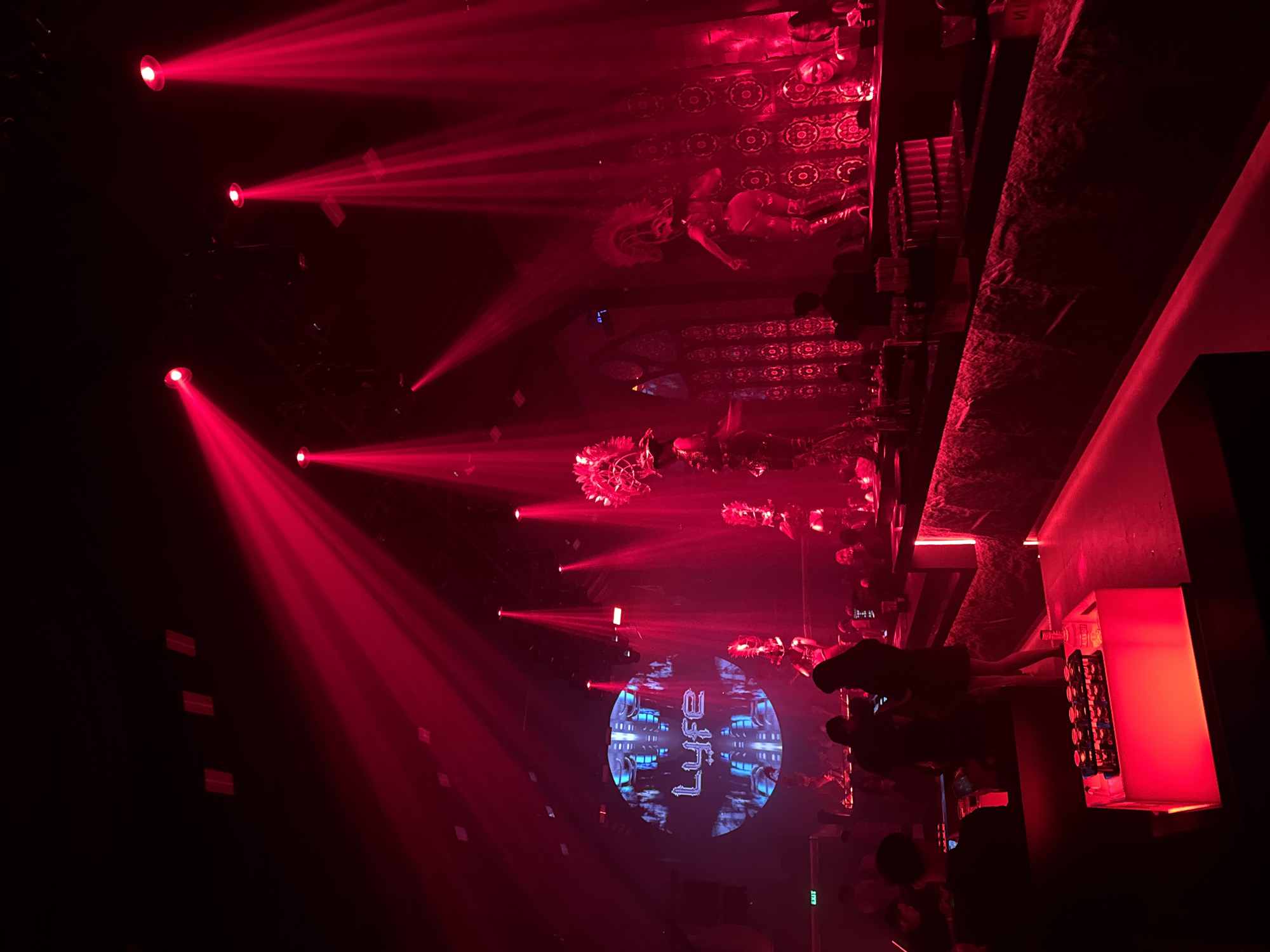 The image depicts the interior of a nightclub in North Kuta, Indonesia, characterized by intense red lighting and a dimly lit atmosphere. Prominently displayed on a large circular screen at the back of the venue is the word "Lyfe" in a stylized, futuristic font, set against a geometric blue and gray background. Several performers, adorned with elaborate headdresses and costumes, are visible on a raised platform, suggesting a live performance or entertainment act. Red spotlights illuminate the space from above, casting beams that cut through the haze. A bar area with a black countertop is present in the foreground, with various bottles and glasses. In front of the bar, there is a luminous red box containing rows of shot glasses, some filled. Several patrons are scattered throughout the nightclub, some standing at the bar, others seated, and some silhouetted in the foreground. An "EXIT" sign is visible on the left side of the image. The overall impression is a vibrant and energetic nightclub environment.
