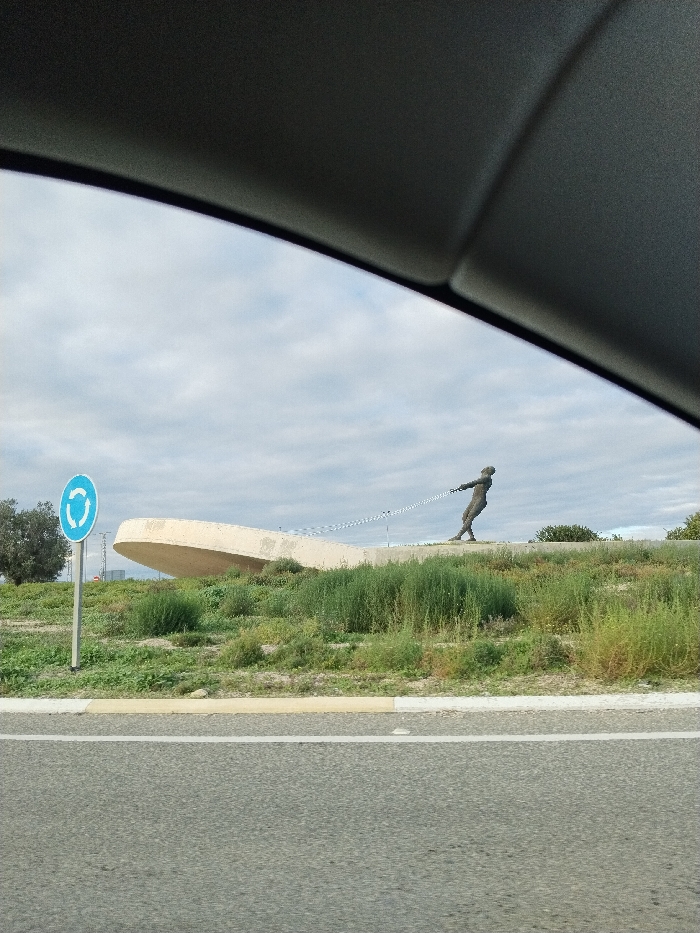 The image, captured from inside a vehicle, displays a public sculpture within a roadside environment under an overcast sky in San Javier, Spain. The upper portion of the frame is occupied by the dark gray interior trim of the vehicle.
In the foreground, a segment of asphalt road is visible, featuring a solid white lane line and a yellow curb to its right. Beyond the road, an unkempt grassy embankment covered with varying heights of green and dry vegetation ascends.
Positioned on this embankment, towards the center-right, is a dark-colored, anthropomorphic sculpture of a person, appearing to be male, in a straining posture. This figure pulls a metallic chain that extends leftwards towards a large, light-colored concrete structure. This structure, resembling a stylized boat hull or wave, is partially embedded in the embankment behind the figure and curves upwards.
To the left of the scene, on a metal pole emerging from the grassy area, a standard blue circular road sign with white arrows indicating a roundabout is visible. The sky is predominantly gray and cloudy. Minimal distant foliage is discernible on the horizon to the far left and right.