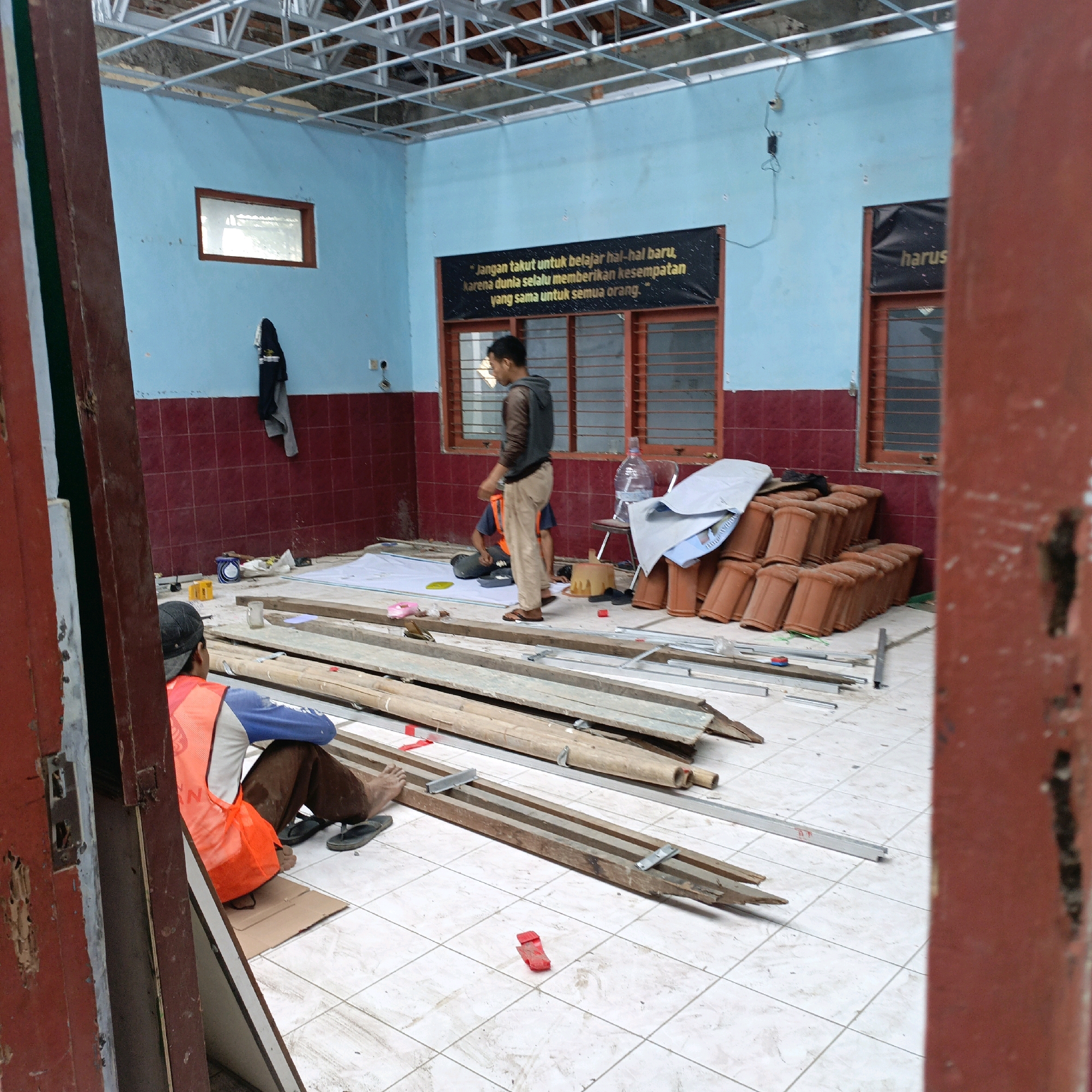 The image depicts an indoor construction site, likely a room under renovation, situated in the city of Salatiga, Indonesia. The room has light blue walls with a red tiled lower section. A metal ceiling grid is visible, indicating ongoing work. Two men are present. One man, wearing an orange safety vest over a blue and white shirt and brown pants, is seated on the floor in the foreground, facing away from the camera. His back is to a pile of wooden planks and metal framing materials. The second man, wearing a gray hoodie and khaki pants, stands near the center of the room, looking downwards. Scattered on the floor are building materials including stacks of terracotta roof tiles, rolls of fabric or sheeting, and various lengths of wood. A few personal items such as a bottle of water and a plastic pink object are also visible. The overall scene suggests a pause in work or preparation for further construction activities.