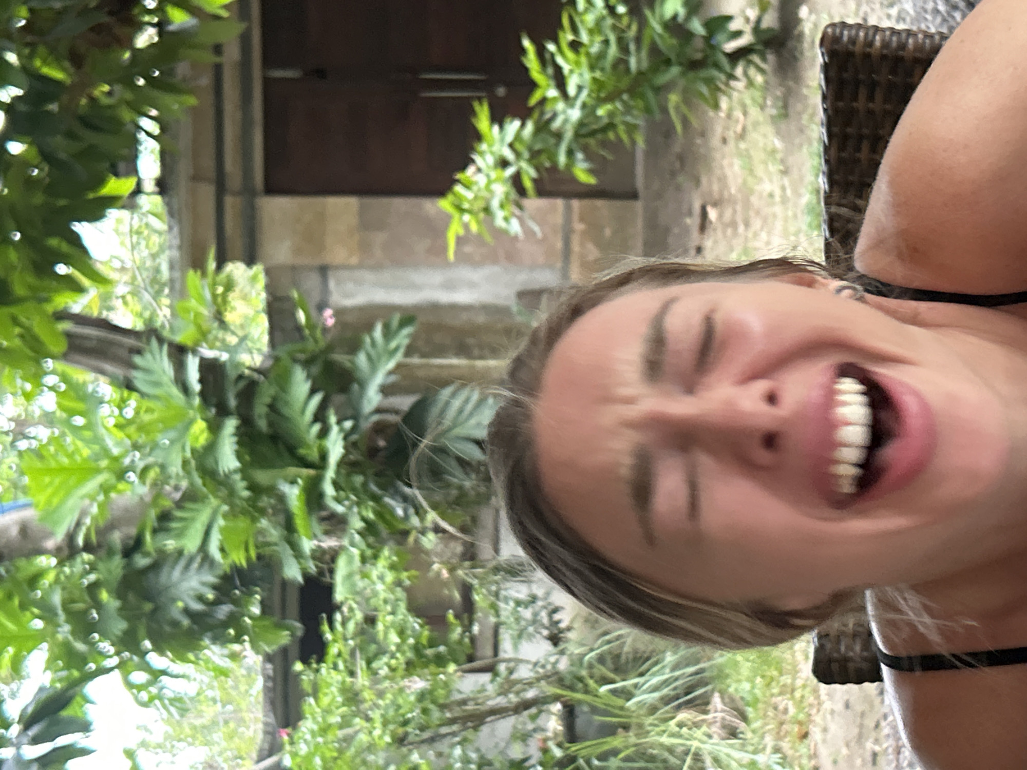 In the city of North Kuta, Indonesia, a woman is captured in a close-up shot, her face contorted in a hearty laugh with her eyes squeezed shut and her mouth wide open, revealing her teeth. She is wearing a black camisole and sits on a dark wicker chair. The background features lush green foliage and a stone building with wooden doors, suggesting an outdoor setting, possibly a garden or patio area. The lighting is bright, and the focus is on the woman's expressive face.
