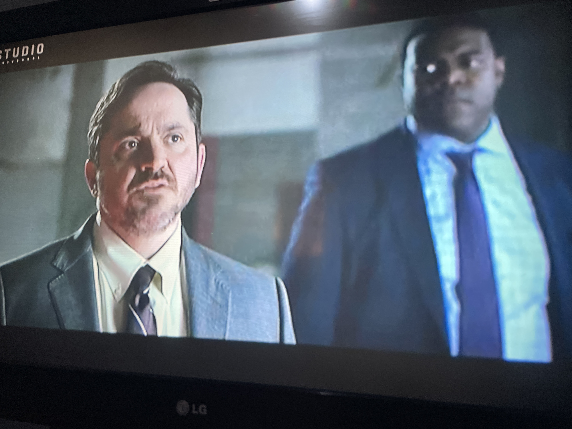 The image shows a close-up of two men in suits, likely from a television screen. The man on the left is in focus, with dark hair, a mustache, and a light-colored shirt with a dark tie. He is wearing a gray suit jacket. The man on the right is out of focus and appears to be wearing a dark suit and a light blue shirt with a darker tie. There is a logo at the top left of the screen that reads "STUDIO" with dots beneath it. There is also an "LG" logo at the bottom center of the screen. The background appears to be an indoor setting with muted colors. There is no discernible location in the city of Jos, Nigeria.