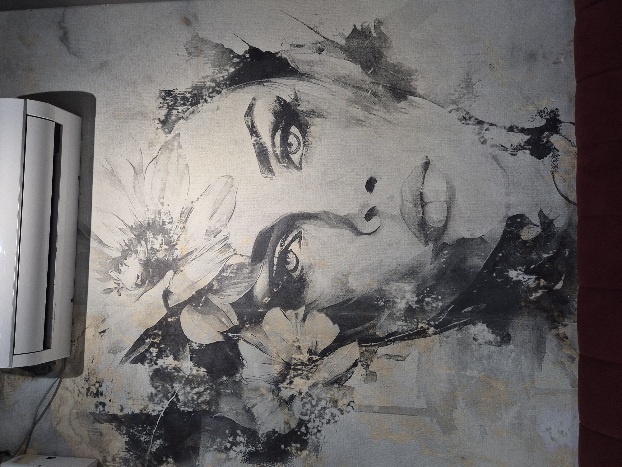 The image depicts a close-up of a monochromatic mural on a textured wall, featuring a stylized portrait of a woman's face adorned with flowers. The artwork utilizes bold brushstrokes and abstract elements, primarily in shades of black, white, and grey, with subtle hints of beige. The woman's features are rendered with dramatic shading, emphasizing her eyes and lips. Surrounding her head are abstract floral motifs, also in black and white, contributing to the overall artistic and somewhat ethereal feel of the piece. The mural is mounted on a wall that appears to be in an indoor setting, possibly a room. Above the mural, a white air conditioning unit is visible, with its vent slightly ajar. The lower portion of the image shows the top edge of a dark red, tufted sofa. The background wall has a textured appearance, with some areas of discoloration or unevenness that contribute to the aesthetic. This mural is located in Kokshetau, Kazakhstan, specifically at the "Chocolat" cafe on Abay Street.