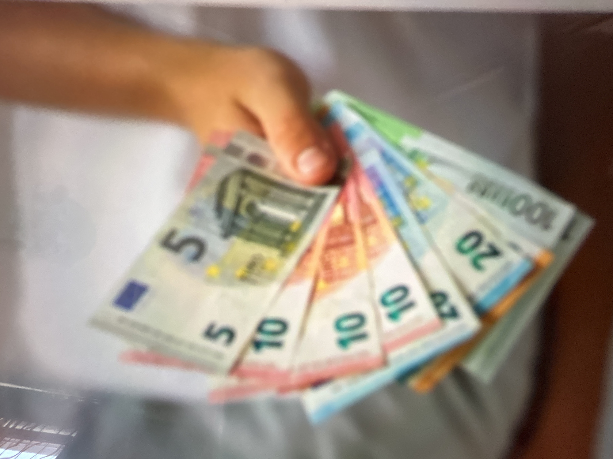 A person's hand is holding a fan of Euro banknotes, fanned out and angled upwards. The banknotes appear to be of various denominations, including 5, 10, 20, and 100 Euros. The image is slightly out of focus, giving it a soft, blurry quality. The background is indistinct, suggesting an indoor setting with soft lighting. There is no identifiable location in the city of Gelendzhik, Russia.