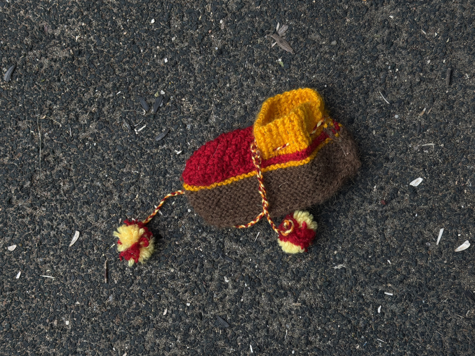 A knitted object, possibly a small hat or a toy, lies on a dark asphalt surface. The object features sections of brown, red, and yellow knit. It has a distinct opening at one end, trimmed with yellow and red stripes, and two braided cords extend from its sides, each ending in a small, fluffy pom-pom made of red and yellow yarn. The asphalt surface is textured and speckled with small gravel and debris, including what appear to be fragments of seeds or plant matter, and small white flecks. The lighting suggests it is daytime, with sunlight casting subtle shadows, though no specific time of day can be definitively determined. The overall scene is simple and static, with the object isolated on the ground. There is no visible text or any indication of people or animals. The context provided indicates the location is Moscow, Russia, which is a factual detail but not visually evident from the image itself. The weather appears to be clear enough for outdoor lighting.