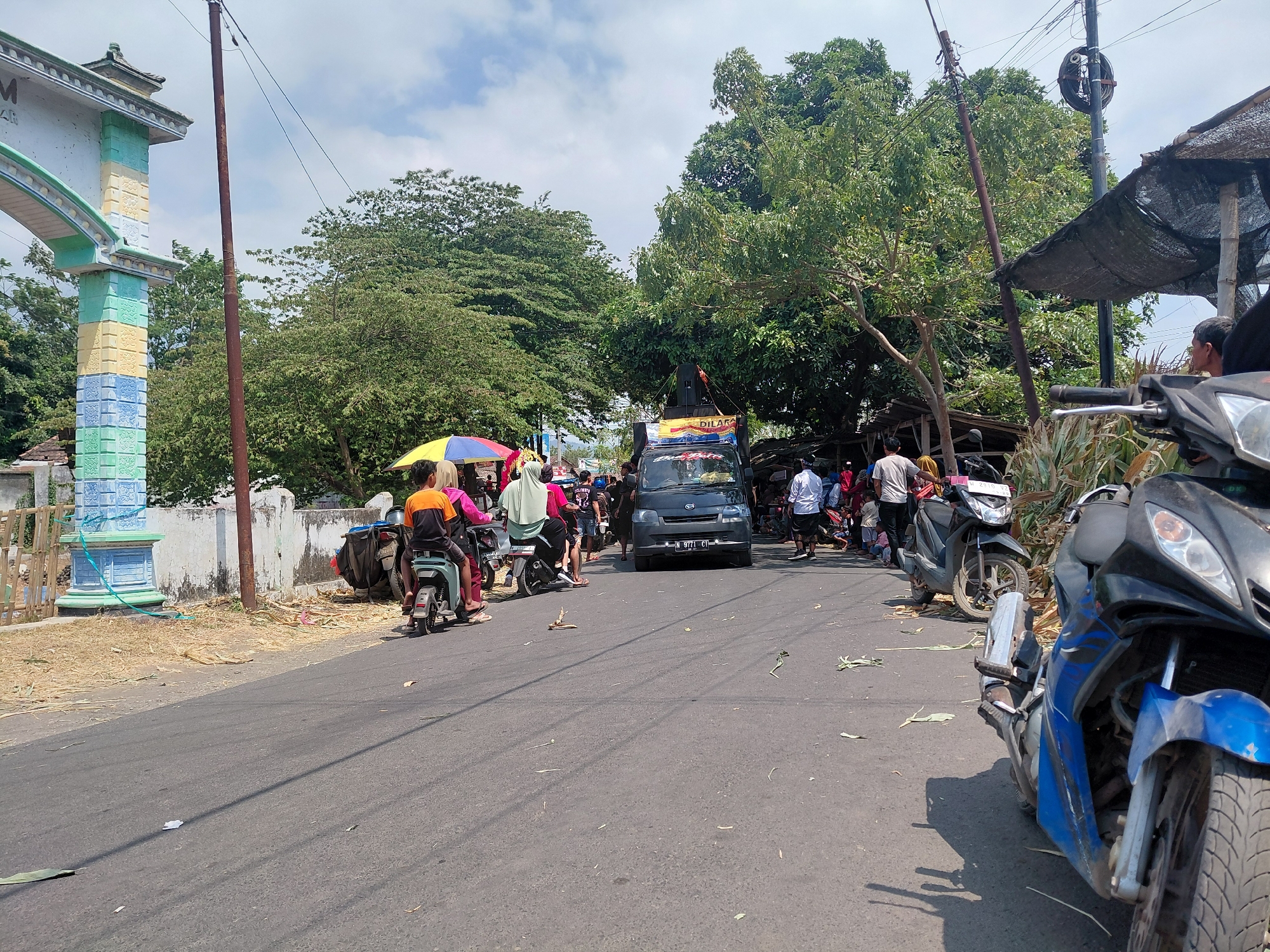 The image captures a street scene in Bimo, Indonesia, on a bright, sunny day with scattered clouds. A main road, likely a local thoroughfare, is the central focus, with vehicles and pedestrians present.
On the left side of the road, a colorful, ornate archway stands, likely marking an entrance to a village or area. It is decorated with pastel colors like teal, yellow, and blue, and features intricate carvings. Adjacent to the archway is a white concrete wall and a short wooden fence. Dry, straw-like vegetation is scattered along the roadside.
The road itself is paved and appears to be the main route for traffic. Several motorcycles are visible, some parked and others in motion. A dark gray van, possibly a commercial vehicle, is driving down the road, displaying a license plate. A smaller, light blue electric scooter is also present, carrying two people.
A group of individuals are gathered near the side of the road, some appearing to be spectators or participants in an event. One person is holding a colorful umbrella, providing shade. Many people are wearing casual clothing. There is a noticeable cluster of people and motorcycles in the middle ground, suggesting some form of gathering or traffic bottleneck.
In the background, lush green trees with dense foliage dominate the landscape, indicating a tropical environment. Some makeshift structures, possibly stalls or shelters with dark fabric coverings, are present on the right side of the road. A power pole with visible wires extends upwards, a common feature in urban and semi-urban areas.
The overall impression is a snapshot of daily life in a Indonesian town, with a mix of traffic, people, and local architecture set against a backdrop of vibrant greenery. The presence of multiple motorcycles and the gathering of people suggest a possibly active or event-filled moment.