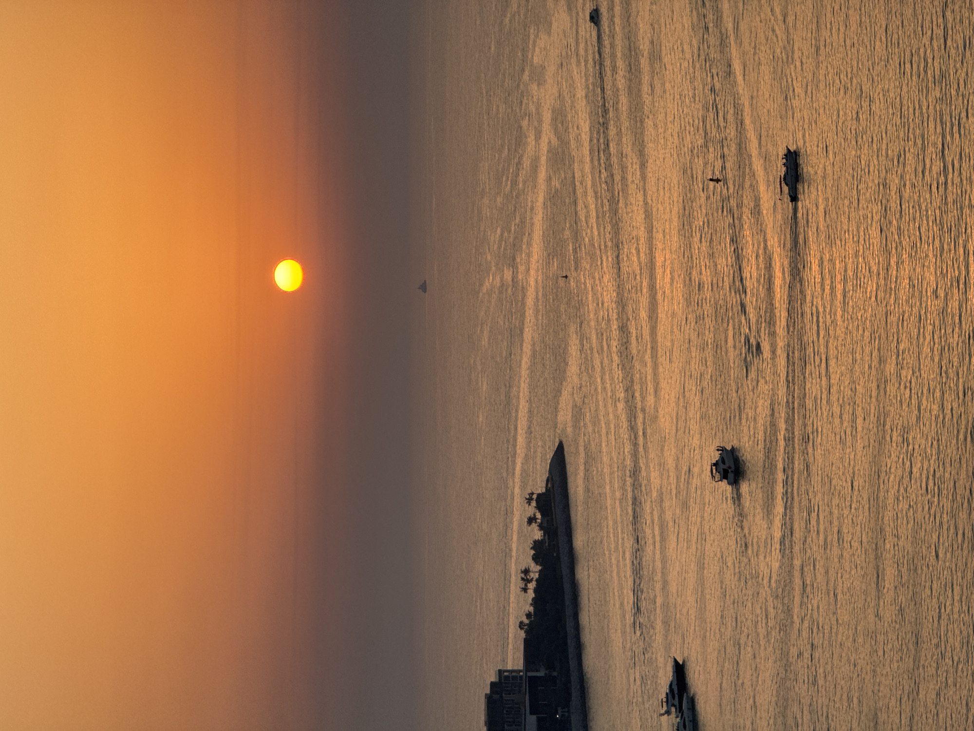 This is a vertical, high-angle photograph of a sunset over the ocean, likely taken from a building in Dubai, United Arab Emirates. The sun, a bright yellow orb, is positioned in the upper left quadrant, casting a warm, orange glow across the sky and reflecting on the rippling water. The ocean fills the majority of the frame, with its surface textured by the setting sun's light, creating streaks of gold and shadow.
Several objects are visible on the water. In the foreground, a dark, elongated structure, possibly a jetty or a breakwater, juts out into the sea from the bottom right. On top of this structure, silhouetted against the water, are palm trees and buildings. Further out on the water, there are several boats of varying sizes. One appears to be a larger vessel, while others are smaller and more agile. Tiny dark specks, possibly birds, are also visible in the sky. A distant, dark shape on the horizon might be another boat or a landmass.
The overall mood is serene and tranquil, characteristic of a sunset over water. The warm colors evoke a sense of peace and the fading of the day. The presence of boats suggests maritime activity, perhaps fishing or recreational trips. The clear sky, with only the sun and a few distant clouds, indicates fair weather. The time of day is clearly sunset, given the low angle of the sun and the golden light. The location cues, such as palm trees and modern buildings, along with the context of Dubai, suggest a coastal setting in the UAE. No visible text is present in the image.