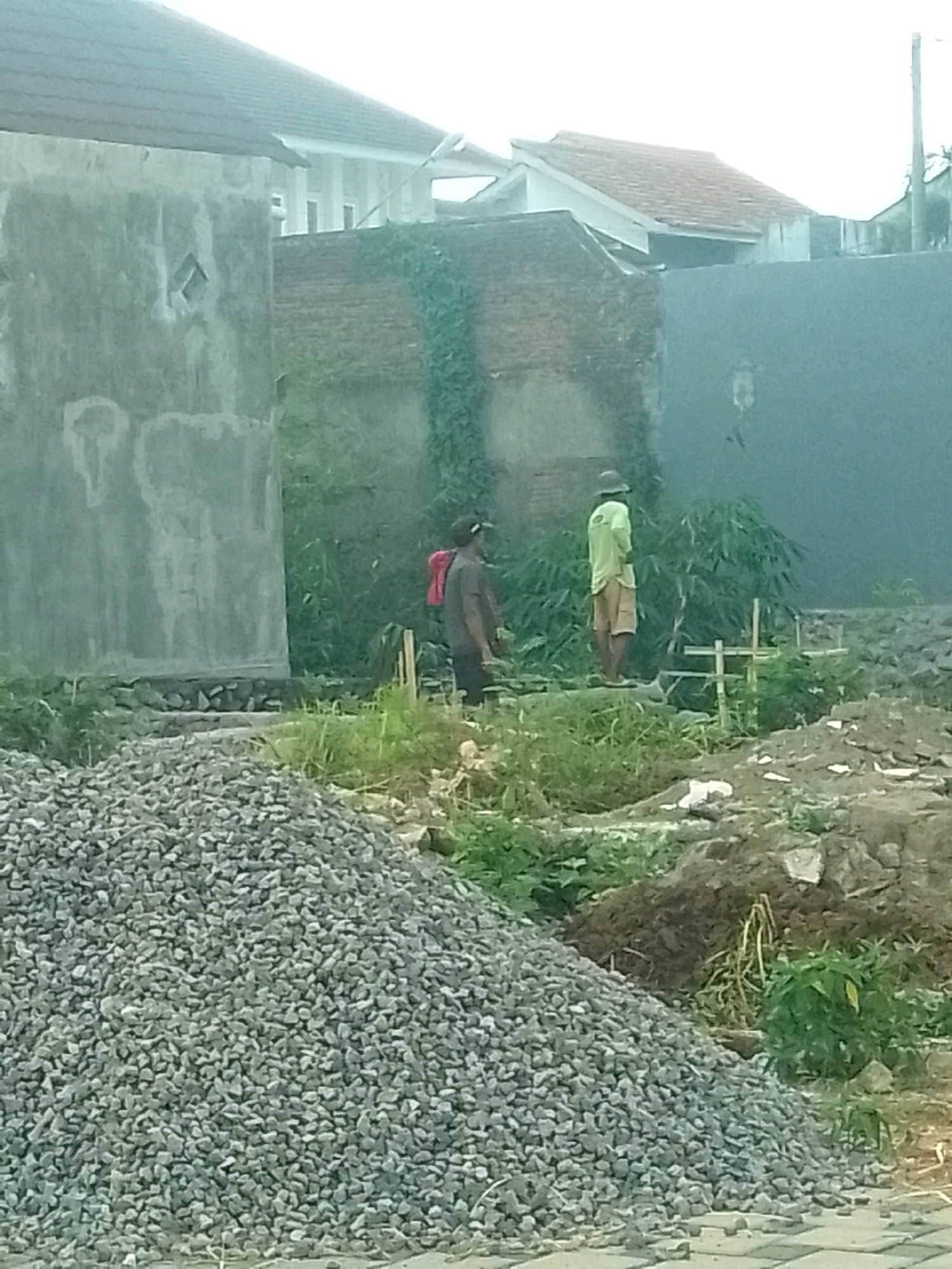 This is a medium-shot, slightly blurred, outdoor photograph of a construction site in Jember, Indonesia. In the foreground, to the left, is a large pile of grey gravel. To the right, the ground is uneven with patches of dirt, rocks, and green vegetation. In the mid-ground, two men are present. The man on the left is wearing a dark shirt and has a red backpack slung over his shoulder. He is facing away from the camera. The man on the right is wearing a light green shirt and light-colored shorts, and a hat. He is standing and facing to the left, away from the camera. There are some thin, light-colored wooden stakes in the ground behind him. In the background, there is a rough, grey concrete wall on the far left, followed by a brick wall covered in green vines. Further back, there is a white building with a tiled roof and several windows, and beyond that, another building with a red tiled roof. The sky is overcast and bright, suggesting it is daytime.