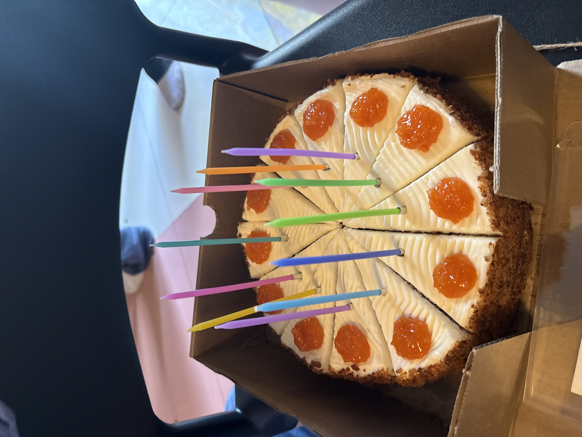 The image shows a round cake in a cardboard box, sitting on what appears to be a black chair. The cake is pre-sliced into eight sections, each topped with a dollop of bright orange jam. Multiple birthday candles in various colors (purple, orange, green, pink, yellow, blue) are stuck into the cake slices. The cake has white frosting and a crumbly edge. Part of someone's legs and feet can be seen in the background, indicating someone is nearby. It's plausible the image was taken indoors, likely for a birthday celebration. There is no definitive information in the image to confirm the location as Saint Petersburg, Russia.