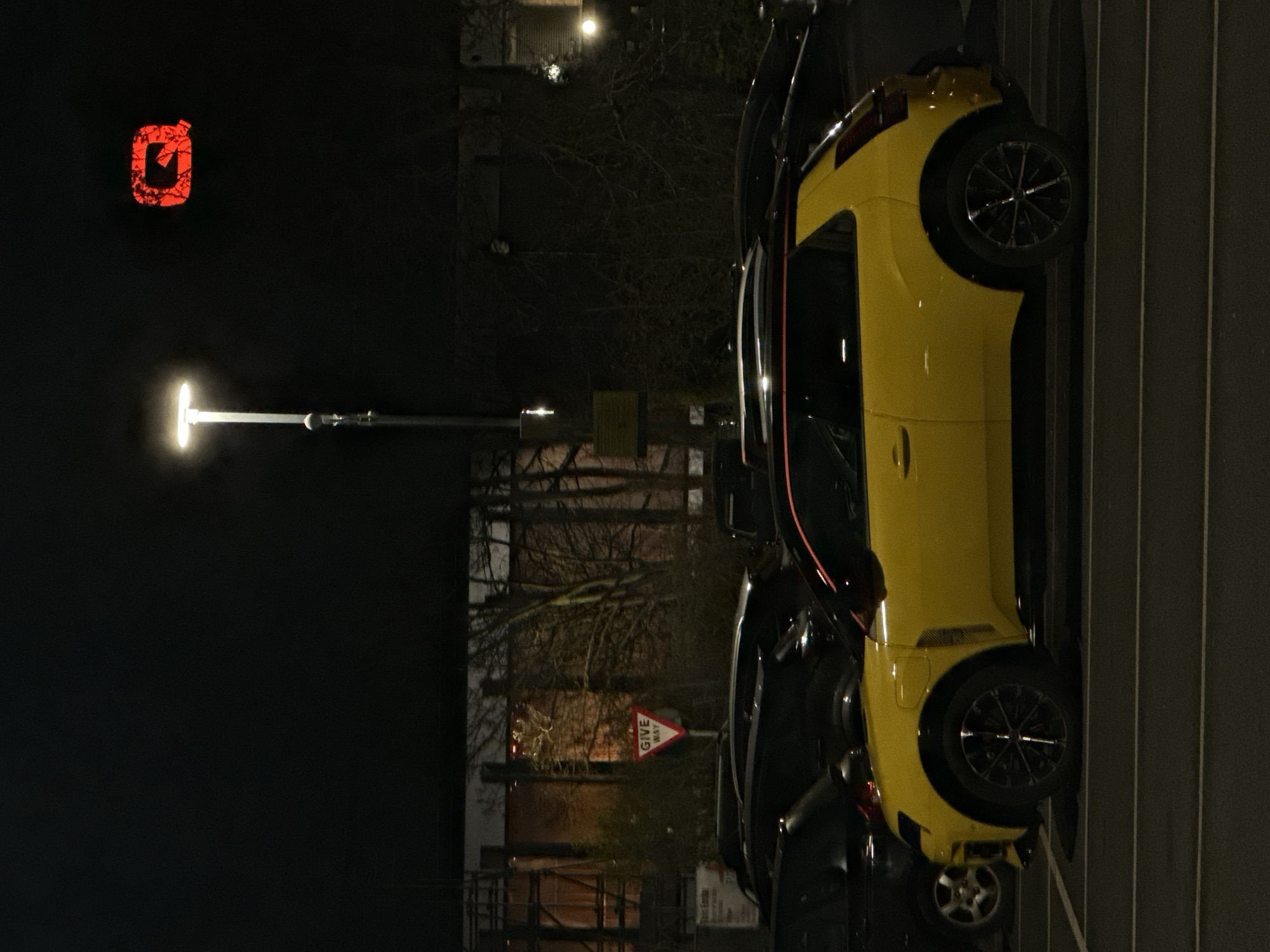 The image depicts a nighttime scene in a car park located near the O2 Arena in Greenwich, London, United Kingdom. In the foreground, a bright yellow modern hatchback, possibly an electric vehicle, is parked facing right, displaying dark multi-spoke wheels and a red trim detail along its roofline. Behind it, several other dark-colored vehicles are parked, partially obscured. A tall streetlamp, emitting a bright white light, stands prominently in the mid-ground. Against the dark sky in the upper right quadrant, a large, illuminated red 'Q' logo is visible. To the left, a white triangular 'GIVE WAY' road sign with a red border is present, positioned amongst bare trees and dark, indistinct building structures in the background. No individuals or dynamic actions are observed in the static scene.