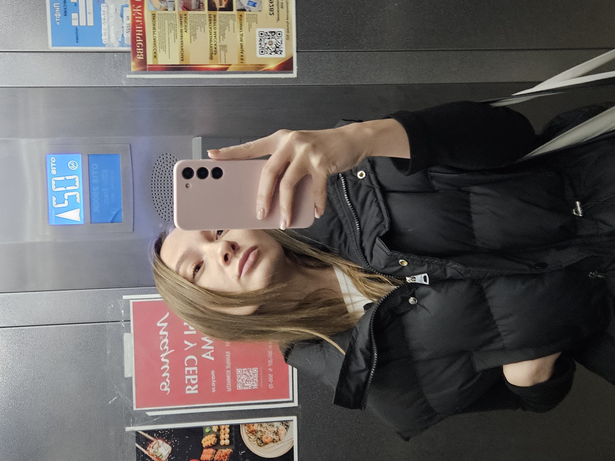 A woman is taking a selfie in an elevator. She is holding a pink smartphone in her right hand. The elevator is decorated with advertisements and information, including a sign with text in Russian and a display showing the number 50. The advertisements visible on the wall include one for a sushi restaurant, indicating the availability of food. The location could be Chaikovsky, Russia due to the Russian advertisements.