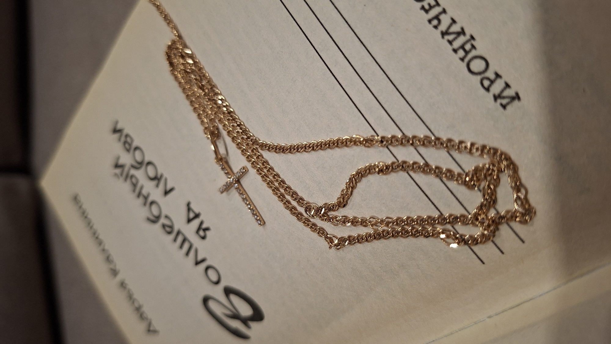 A gold necklace with a cross pendant is lying on a book. The book's pages are visible and show musical notation with lines and some text in a Cyrillic alphabet. The text seems to reference "love." Based on the Cyrillic alphabet, it is likely that the book is in Russian. The environment appears indoors. No specific location is indicated and nothing indicates Chaikovsky, Russia.