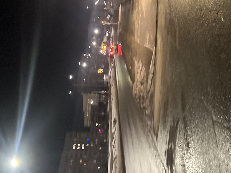 This image depicts a nighttime street scene in Yelabuga, Russia, under artificial illumination. The environment suggests a winter setting, with snow and slush present on the ground, roadside, and median strip. The asphalt road surface appears wet and reflective, indicating either recent precipitation or melting snow.
In the background, multiple multi-story buildings, likely residential, are visible, with some illuminated windows suggesting occupancy. Streetlights line the road, casting bright flares into the upper left of the frame. A decorative, illuminated yellow-orange spherical object, possibly a festive ornament, is visible on a pole in the middle ground, right of center.
A red passenger vehicle is positioned in the middle ground, its rear facing the viewer, with its bright red taillights illuminated and reflecting on the wet road surface. No individuals are visible in the scene. The overall image exhibits motion blur and low-light characteristics, particularly in the foreground and distant background.