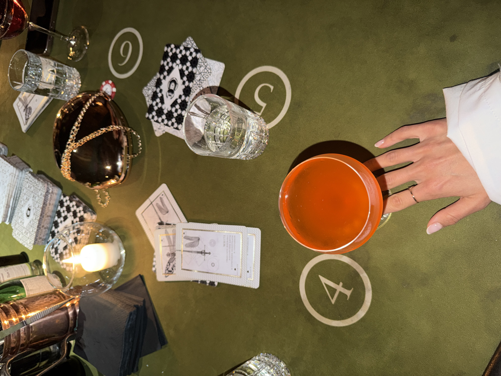 The image captures a tabletop setting, possibly in Saint Petersburg, Russia, characterized by a green felt surface that appears to be a card table. Scattered across the surface are several objects: a lit candle in a glass holder, a stack of black napkins, tarot cards, a metallic spherical bag with a gold chain, two glasses of liquid (one clear, one amber-colored), a deck of cards with a black and white pattern, and a wine glass filled with red liquid. White circles with numbers (4 and 5) are visible on the green surface, suggesting a gaming or divination context. A human hand, adorned with rings, is reaching towards the amber drink. The lighting suggests an indoor, possibly dimly lit environment, with reflections visible on the metallic bag and glassware.