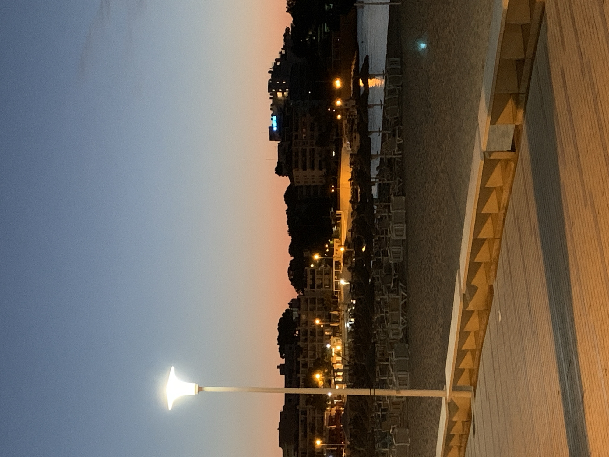 This image depicts a twilight scene on a beach promenade in Palmanova, Spain. In the foreground, a wooden walkway with a low, segmented bench runs horizontally across the frame. Beyond the walkway is a sandy beach with rows of empty sun loungers and parasols, indicating the late hour. The sea is visible in the background, reflecting the lights from the buildings. The cityscape of Palmanova is silhouetted against a gradient sky of deep blue to soft orange, suggesting dusk or dawn. Several buildings, including hotels or apartments, are lit up, with some lights creating bright points. A tall lamppost with a lit, modern fixture stands prominently on the beach. The overall atmosphere is serene and quiet, characteristic of a tourist resort during the transition from day to night.