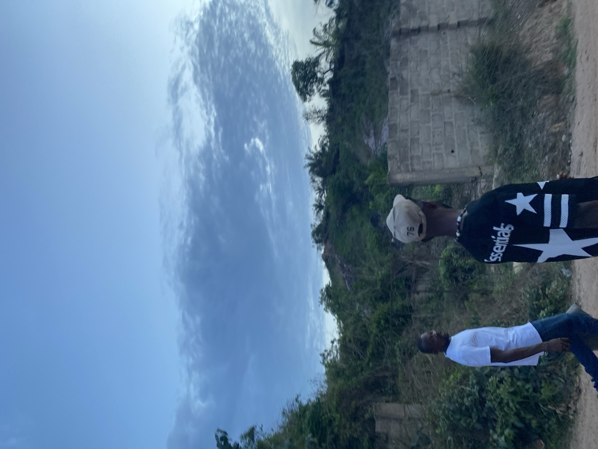 The image shows two men in an outdoor setting in Achara, Nigeria. One man is wearing a white t-shirt and blue jeans. The other man is wearing a black shirt with a white star and the word "Essentials" on it, and a baseball cap with the number "76" on it. The background includes a partly cloudy sky, vegetation, and a brick structure.