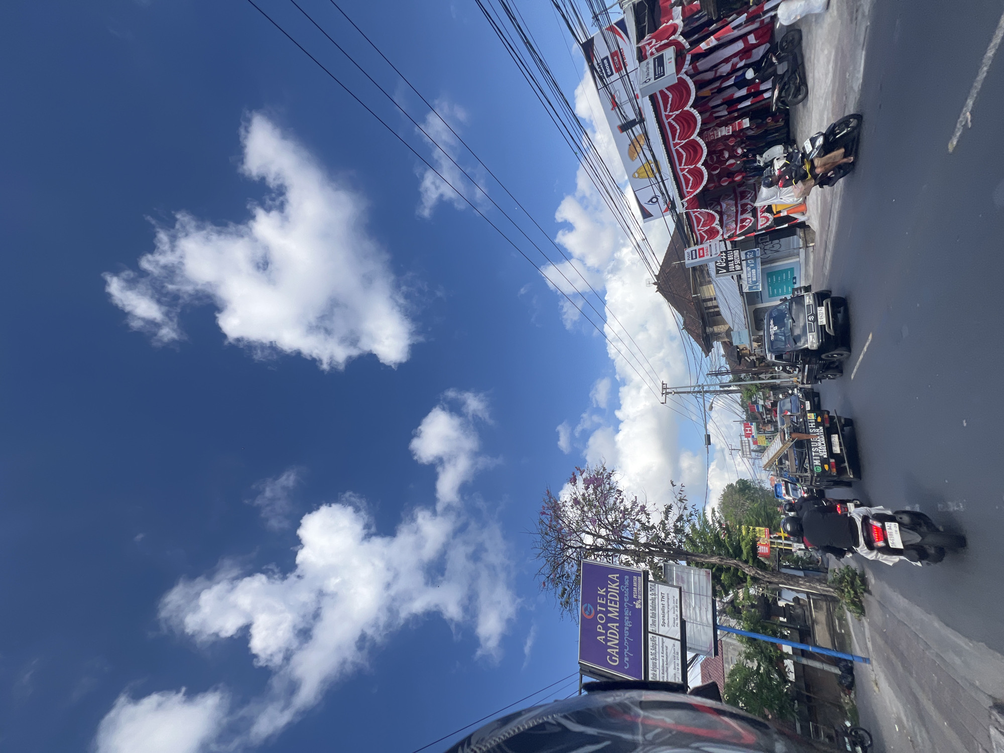 The image depicts a street scene in Denpasar, Indonesia, under a bright blue sky with scattered white clouds. The perspective is from the viewpoint of someone riding a motorcycle. On the left side of the road, a sign for "APOTEK GANDA MEDIKA" is visible, with smaller text indicating services and contact information. A tree with pink blossoms stands near the sign.
The road is paved and appears to be a main thoroughfare with traffic. Several vehicles are present, including motorcycles and a small black pickup truck. The pickup truck has "MITSUBISHI KANGKANG SELAMAT" written on its side and a ladder strapped to its roof. People are riding on the motorcycles, most wearing helmets. One motorcyclist in the foreground is wearing a black jacket and white pants, and their motorcycle has a license plate that reads "DK 6386 AAZ". Another motorcyclist is wearing a yellow jacket and shorts.
On the right side of the street, buildings are visible, with awnings and decorations, including red and white banners, suggesting a commercial area. One building has signs that say "PUSAT" and "GROSIR". Another sign says "JUAL BELI HP SECOND". The scene is bustling with activity, characteristic of a busy urban environment. Electrical wires are strung across the sky, typical of urban infrastructure. The overall impression is a snapshot of daily life on a sunny day in Denpasar.