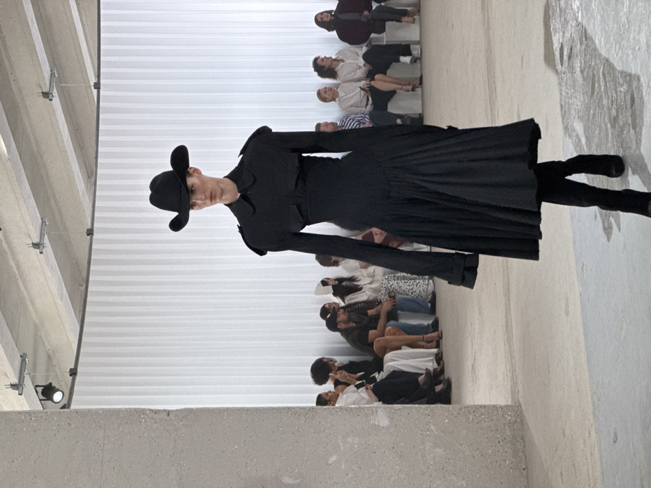 An indoor fashion show is depicted within a modern, minimalist event space in Paris, France. A tall, slender model, centrally positioned and walking towards the viewer, wears a monochromatic black ensemble consisting of a wide-brimmed cowboy-style hat, a long-sleeved collared top with structured shoulders and chest pockets, a cinched waist, and a voluminous pleated midi-length skirt, paired with black boots. To the left and receding into the background, a seated audience of approximately 10-15 individuals observes the runway. They are dressed in varied casual to semi-formal attire, with some holding mobile phones. The venue features a light-colored concrete floor, a curved backdrop of vertical white ridged panels, and an exposed concrete ceiling with visible track lighting fixtures. A prominent, textured concrete pillar stands on the left side of the frame.