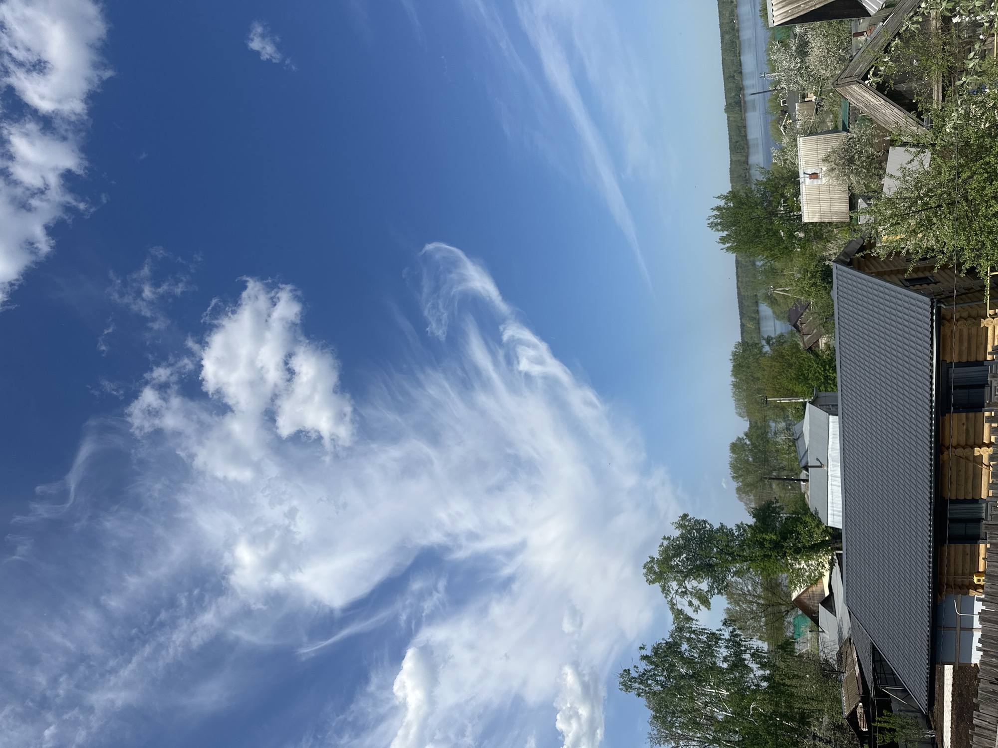 The image shows a rural area, likely in Chaikovsky, Russia. The foreground features a blue sky with wispy clouds, including some cirrus formations. The right side of the image depicts a landscape with several houses and trees. The houses have various roof types, including gray metal roofing. A river is visible in the background, lined with trees along its banks. The scene seems to be shot during the daytime with clear weather conditions.