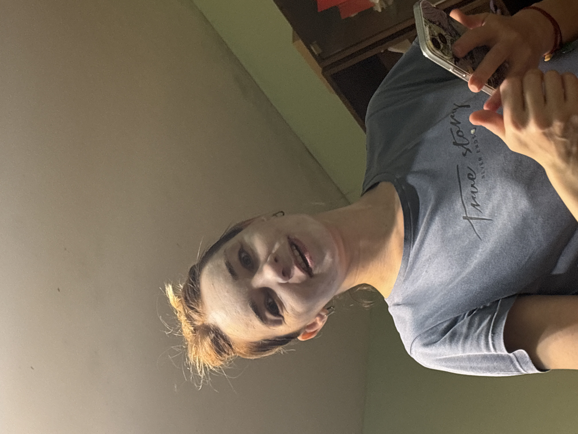 The image depicts a woman holding a phone. She is wearing a blue t-shirt with text, and appears to be in an indoor environment. The location in the city of Unknown, Russia, cannot be determined from the image.