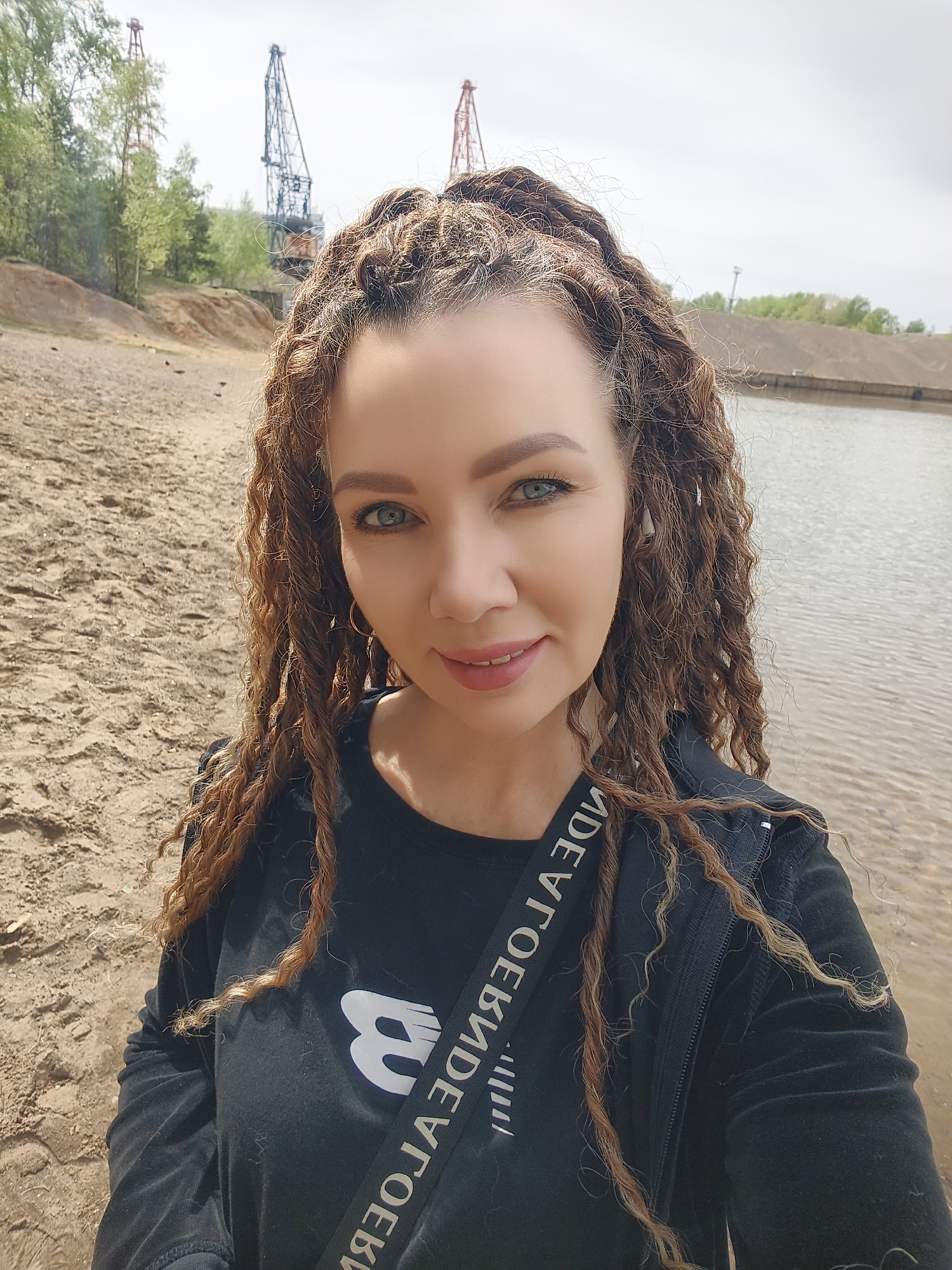 A woman stands on a sandy shore next to a body of water. She has long, curly hair, green eyes, and is wearing a black shirt and jacket with a strap that has text in reverse. Behind her, there are industrial cranes and trees. The location is in the city of Unknown, Unknown.