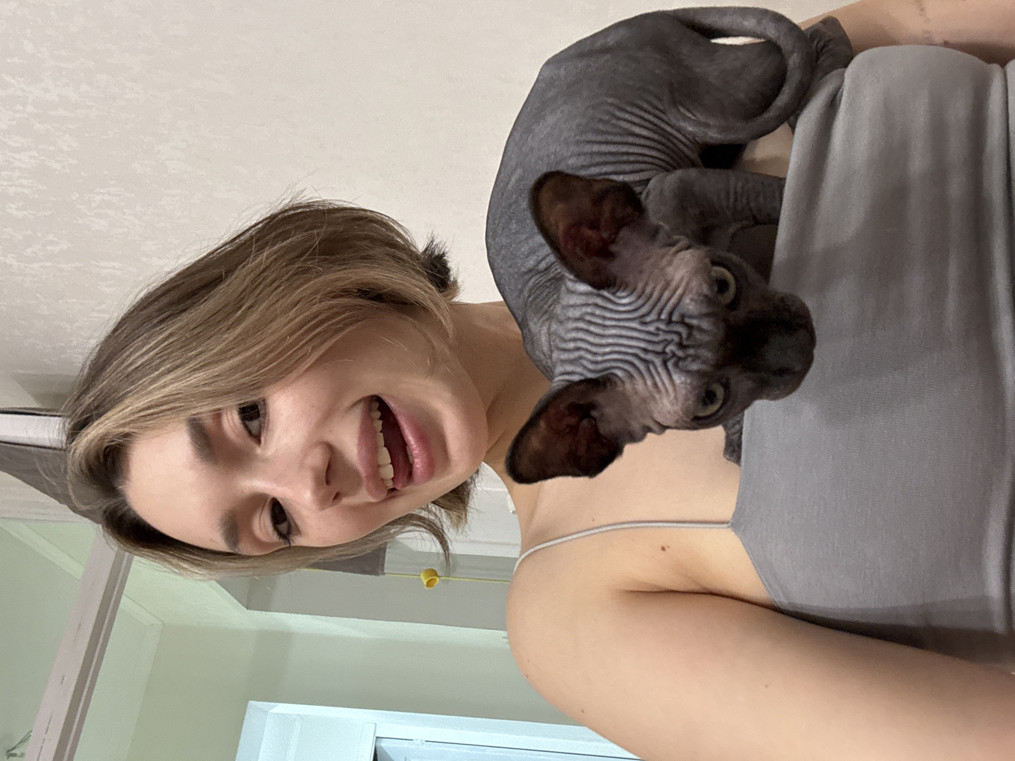 A young woman with short, blonde hair and a playful expression, sticking her tongue out, holds a dark-colored Sphynx cat on her shoulder. She is wearing a grey tank top. The cat is looking directly at the camera with a curious expression. The background appears to be an indoor setting with neutral-colored walls and a doorway visible on the left. The lighting is soft and natural.