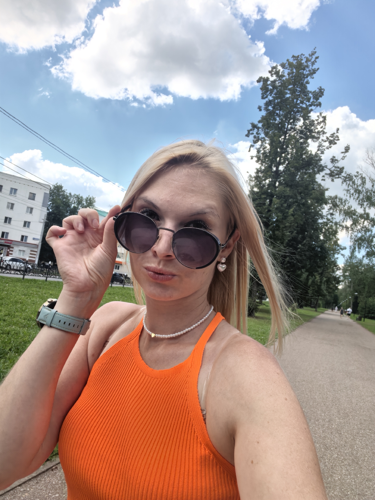 A blonde woman wearing sunglasses, an orange halter top, a pearl necklace with a heart pendant, and a light blue watch is taking a selfie outdoors. She is holding the edge of her sunglasses with her right hand. She appears to be walking on a paved path in a park, with a grassy area and trees to her left and right. In the background, there is a building and a blue sky with white clouds. The scene is set in the city of Sterlitamak, Russia, as indicated by the building's architecture and Russian signage visible on it.
