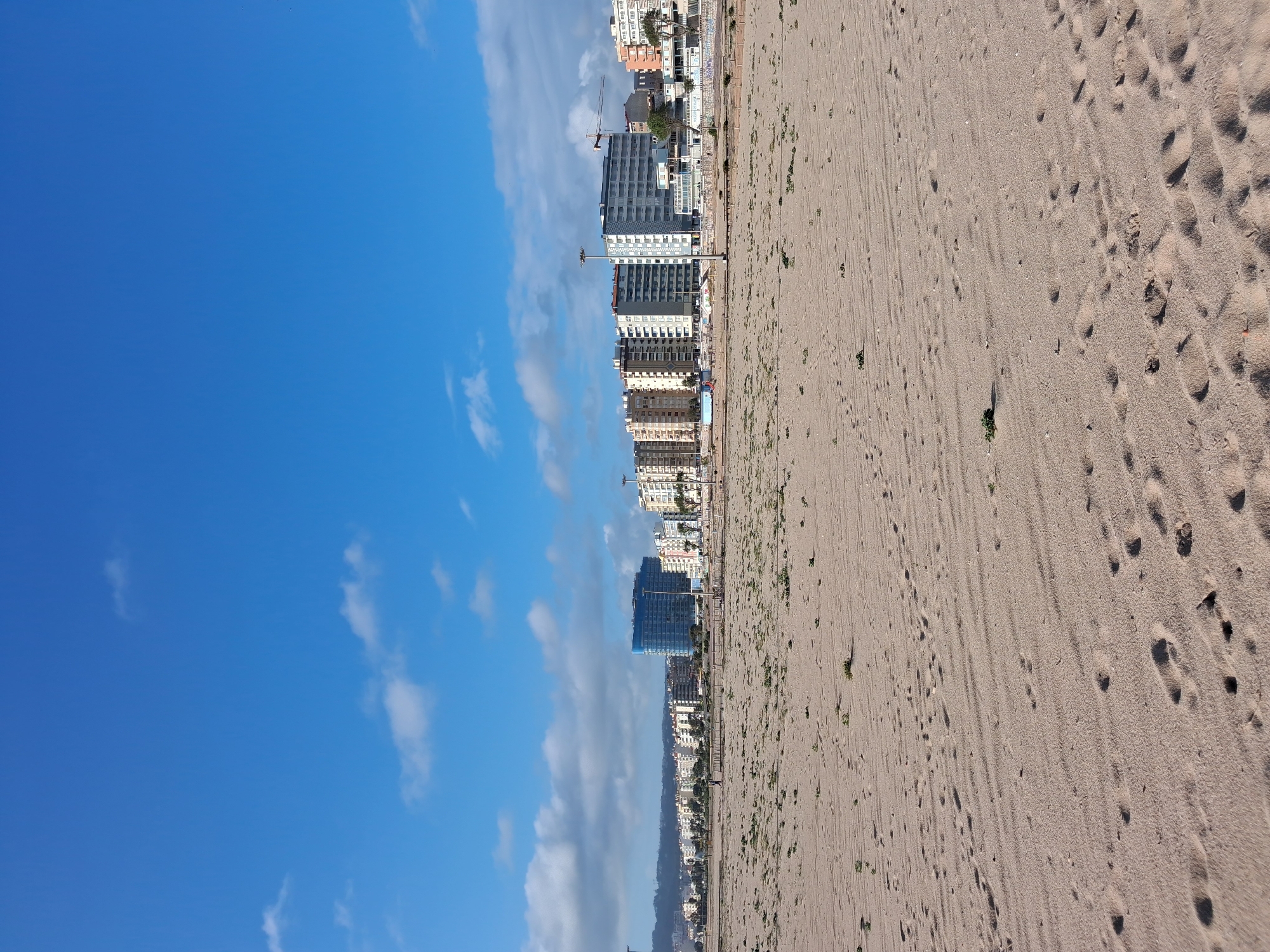 The image shows a beach scene in Figueira da Foz, Portugal. The scene is divided vertically into thirds. The left third is a clear blue sky with sparse white clouds. The central third contains a line of high-rise buildings along the coast. The right third is a sandy beach, marked with various footprints and the occasional clump of vegetation. The image is oriented in a portrait format.