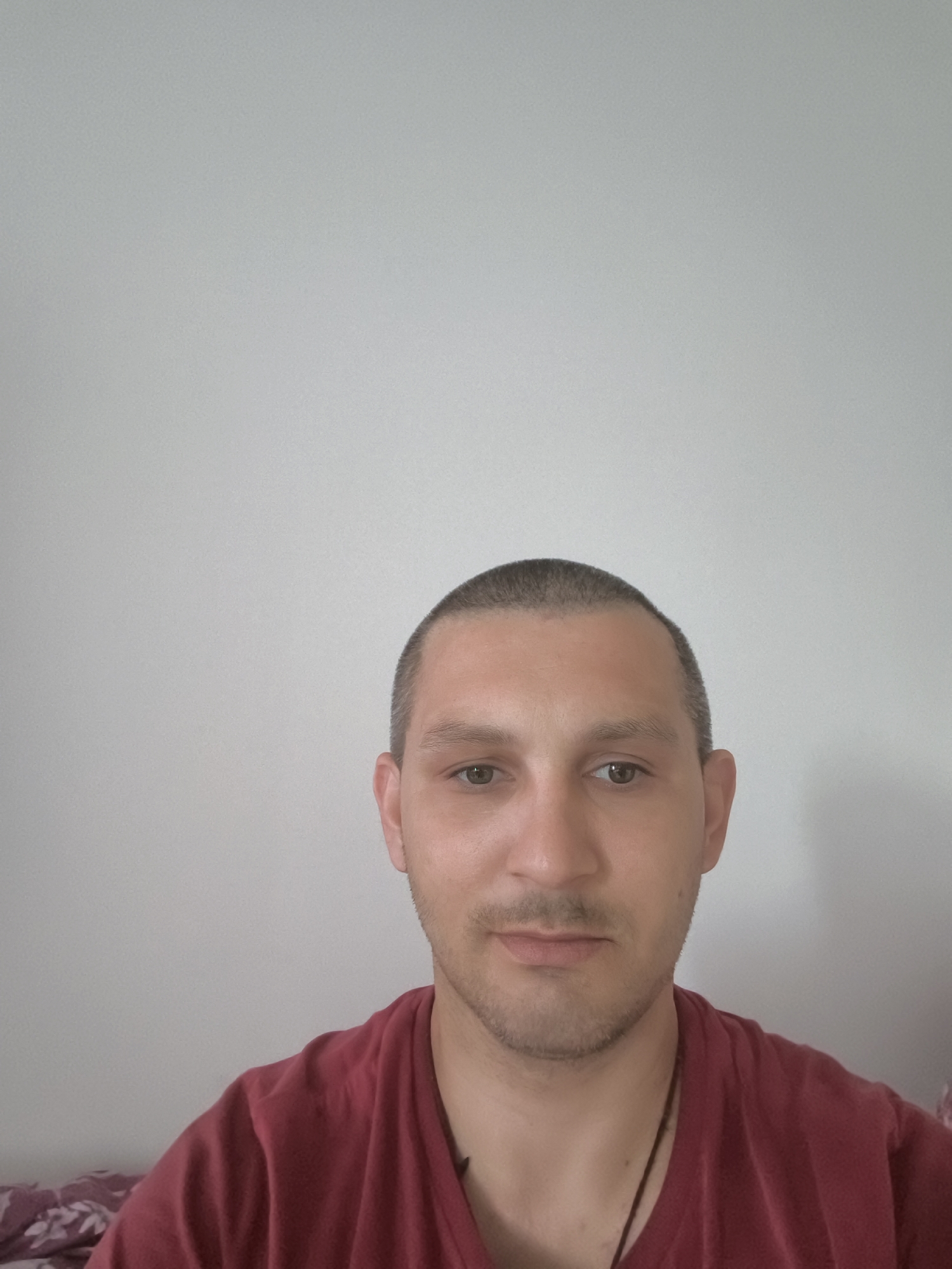 A portrait of a man with a shaved head and brown eyes, wearing a maroon t-shirt. He has a light beard and is looking directly at the camera. The background is a plain, light-colored wall. The scene appears to be indoors, possibly in a home. There is no indication of the location being in the city of Wolgast, Germany.