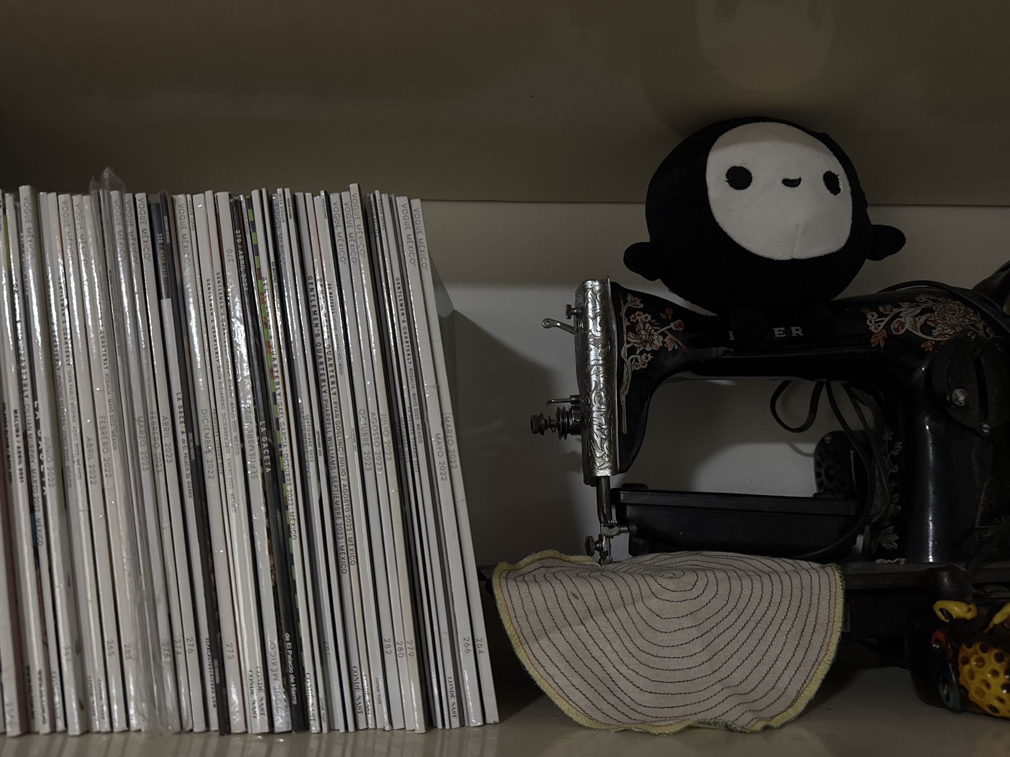 A shelf contains a collection of magazines, a vintage Singer sewing machine, and a plushie. The magazines, primarily titled "Vogue Mexico" and "Gentlemen's Quarterly," are arranged neatly on the left side of the shelf. A black-and-white plush toy with a simple face sits atop the sewing machine. The sewing machine, an antique Singer model, is adorned with floral patterns. A circular piece of fabric with concentric stitch patterns is draped over the machine. A decorative ceramic piece with yellow and brown colors is present to the right of the sewing machine. The shelf appears to be inside a building in Mexico City, Mexico.