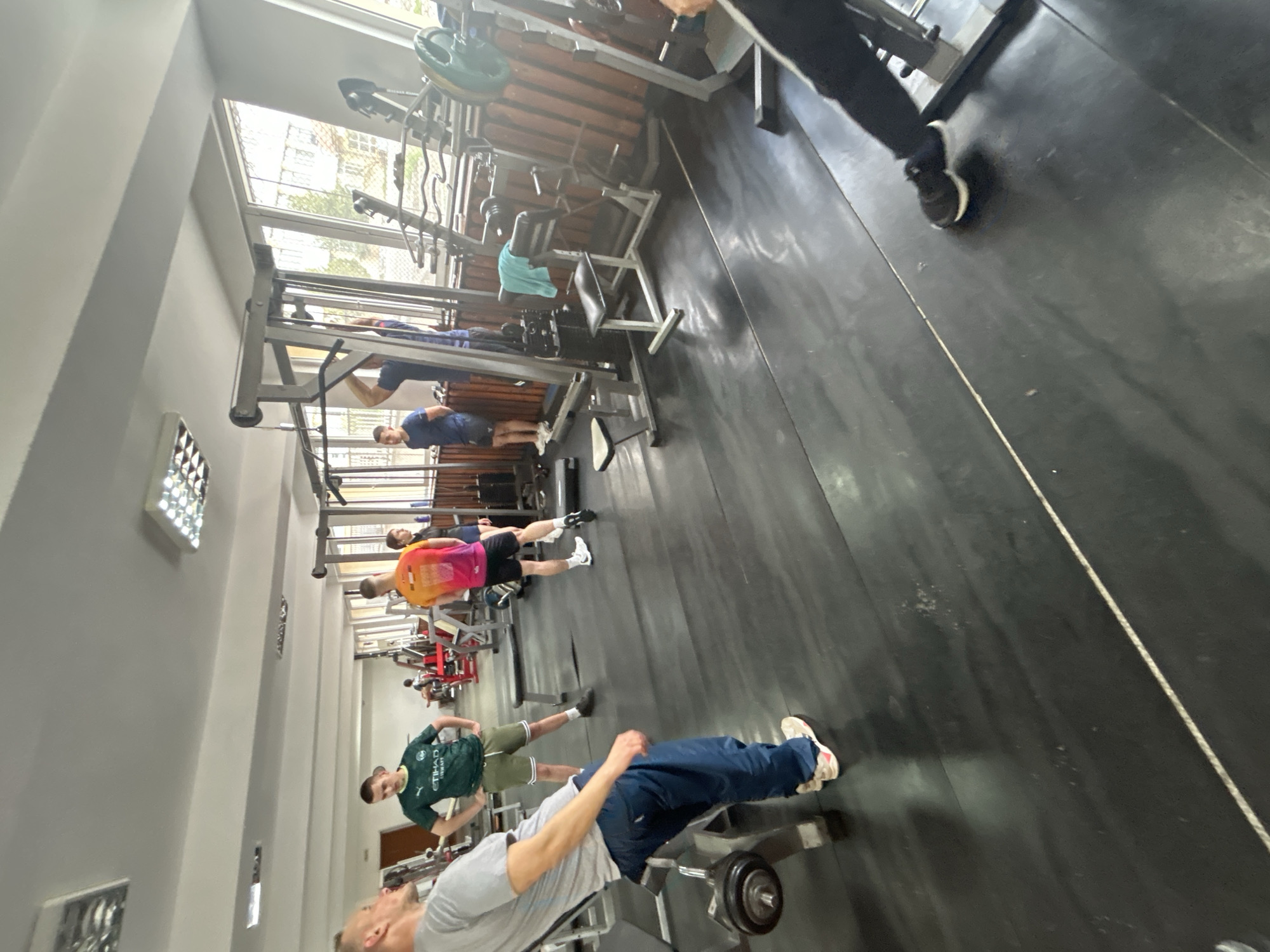 The image depicts the interior of a gym. Several people are present, engaged in various exercises. Equipment includes weight machines, benches, and free weights. The location is likely Zlatograd, Bulgaria.
