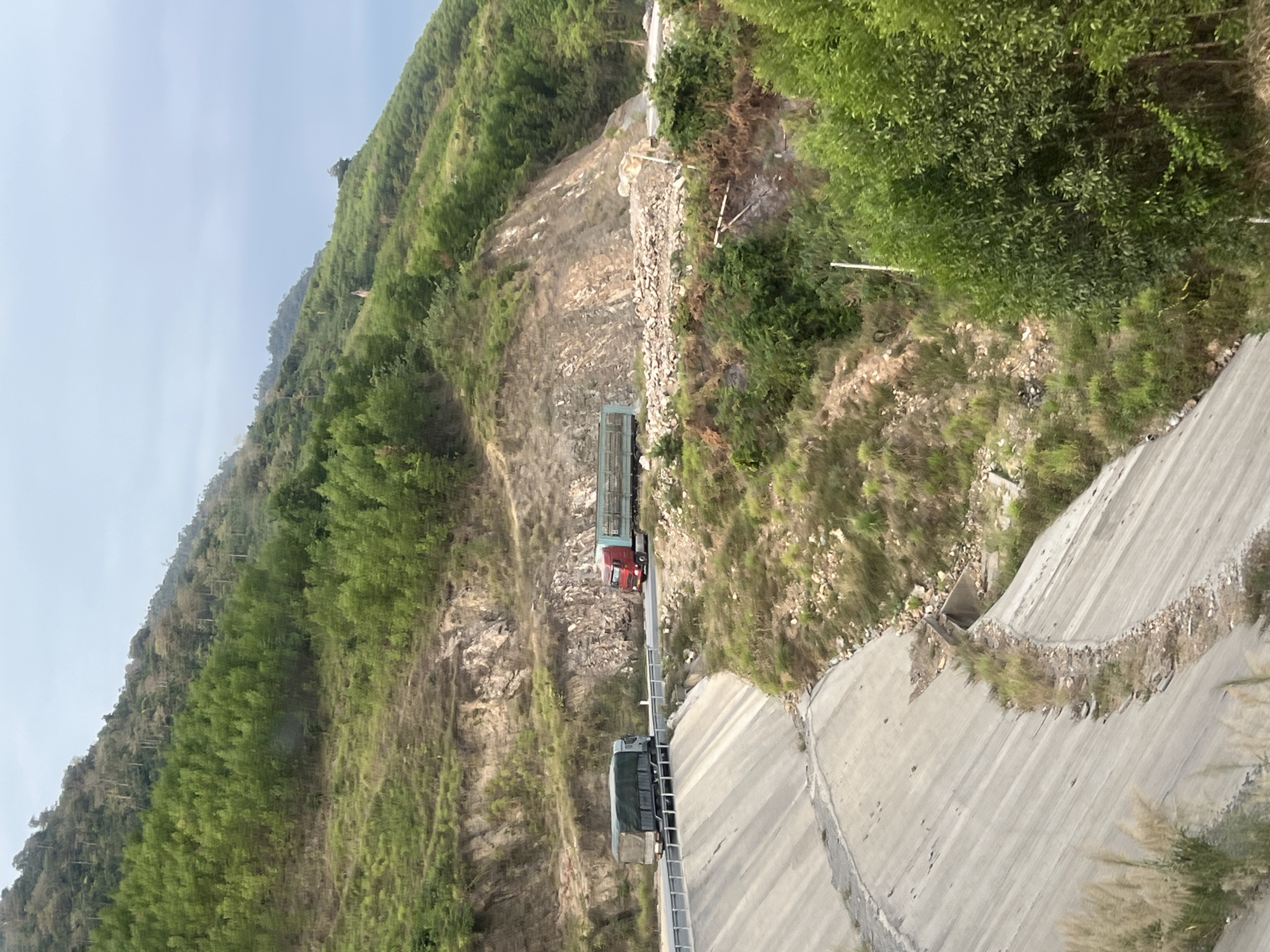 The image depicts a mountainous landscape in Aê Thi, Vietnam, showcasing a road carved into the hillside. A red semi-truck is parked on the road, while a grey semi-truck is further down the road. The roadside features rocky terrain and vegetation. Concrete infrastructure flanks the road, including what appears to be drainage systems. The backdrop is a steep, tree-covered mountain, and the sky is overcast.
