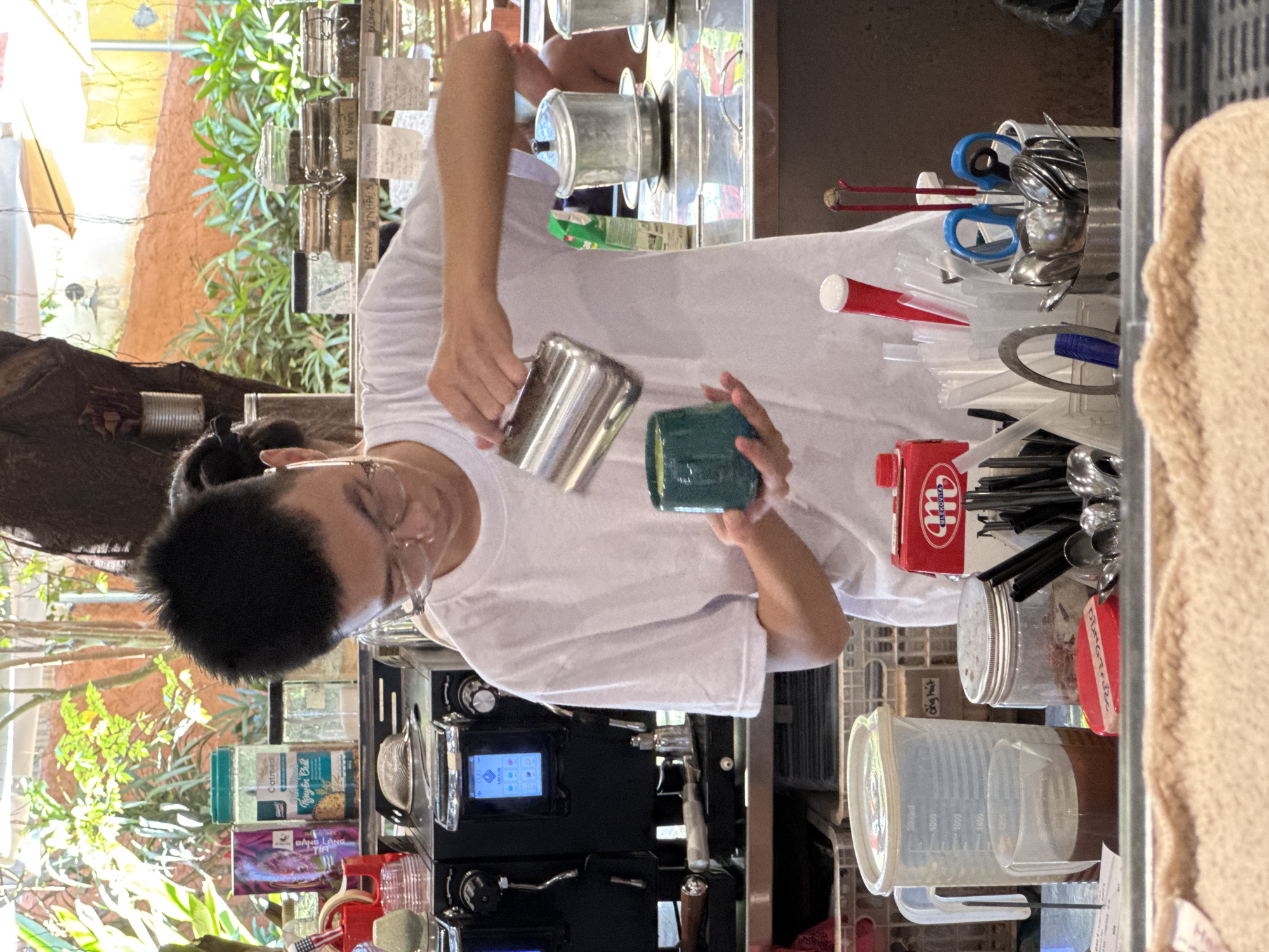 The image depicts a young person with short, dark hair and glasses, wearing a white t-shirt, standing behind a counter at a coffee shop in Nha Trang Ward, Vietnam. The person is holding a metal pitcher and pouring liquid into a teal-colored ceramic cup. In the background, there are several items, including an espresso machine, various containers of coffee beans or other dry goods, and a large clear pitcher filled with a dark liquid. The setting appears to be an outdoor or semi-outdoor area, with sunlight filtering through foliage and an awning visible in the upper portion of the frame. The counter is organized with various coffee-making accessories such as straws, spoons, and a pair of blue-handled scissors. The overall scene suggests a barista preparing a drink in a casual, possibly tropical, environment.
