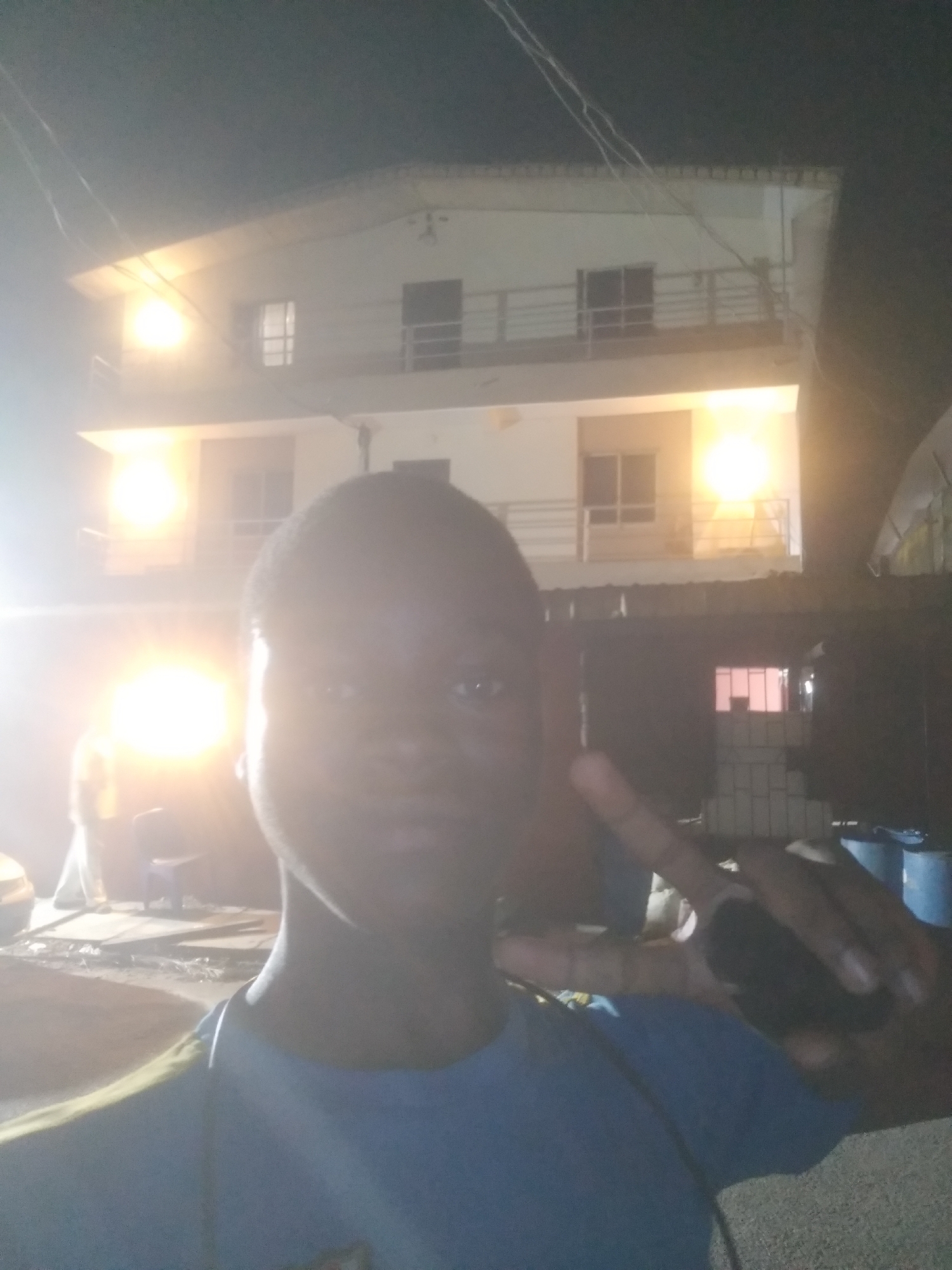 A nighttime selfie captures a young person in the foreground, outdoors in Shangisha, Nigeria. The subject, a person with dark skin, appears to be a teenager or young adult, wearing a blue t-shirt with a yellow or green trim visible on the left shoulder. Their face is largely obscured by intense artificial light and lens flare, making clear facial features indistinct. The person's right hand is raised, making a peace sign gesture.
In the background, a two-to-three-story building is illuminated by several bright light sources, which also contribute to the significant lens flare seen in the image. The building features balconies with metal railings and multiple windows, some of which show interior lighting. The sky above is dark, confirming the nighttime setting.
To the lower right of the building, several blue barrels or drums are visible. On the lower left, some indistinct objects, possibly including a chair, are present. Overhead, faint power lines stretch across the dark sky. The scene is dominated by the strong artificial lighting, creating a high-contrast image. No discernible text is visible.