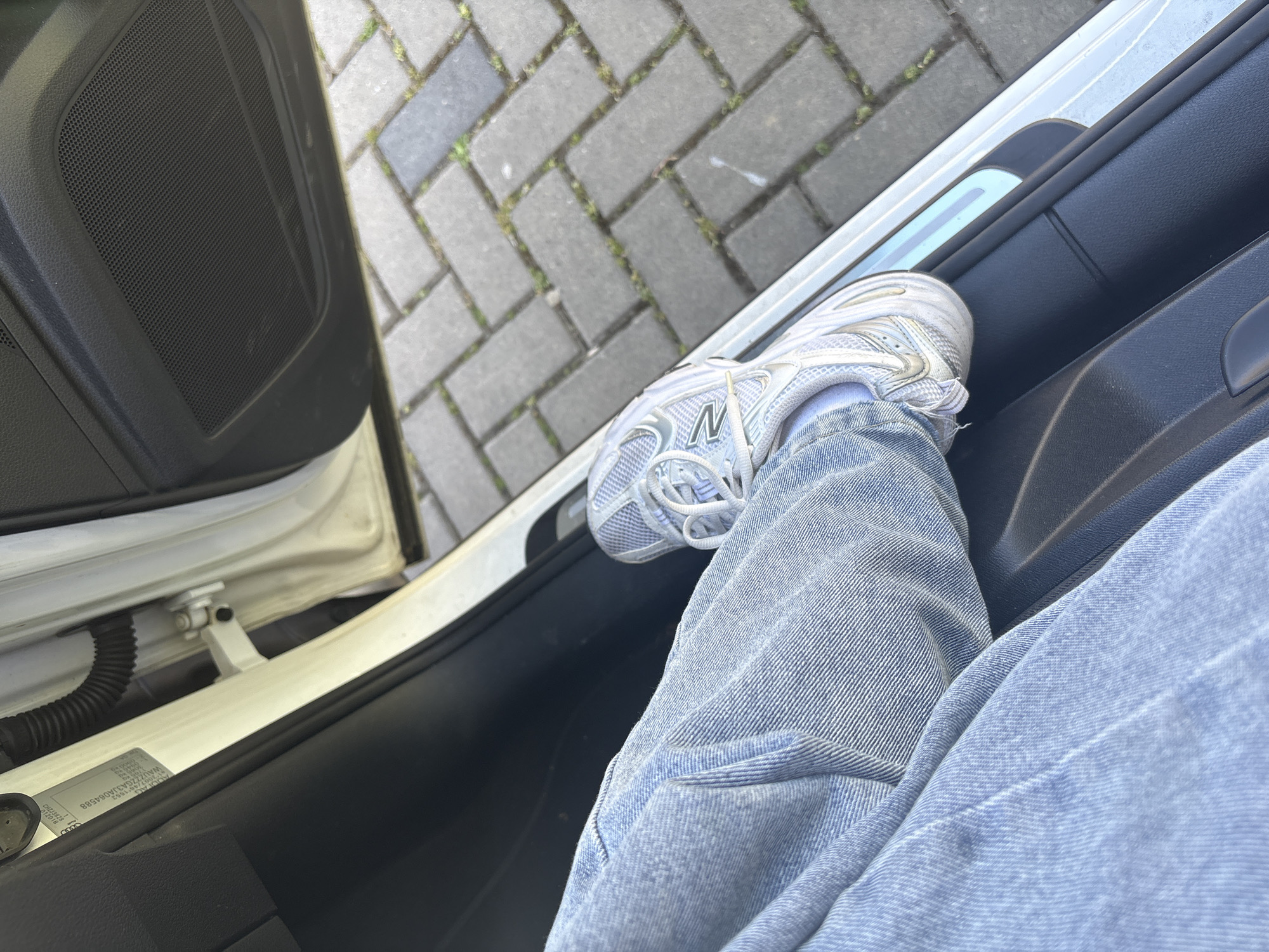 A photograph taken from inside a car shows a person's leg resting on the car door. The person is wearing light blue jeans and a white sneaker, specifically a New Balance 530 model. The car door is open, revealing a view of a brick sidewalk or street outside. A car door hinge and an information label are visible on the car frame. The location is inferred to be in Wassenberg, Germany based on the prompt's requirements.