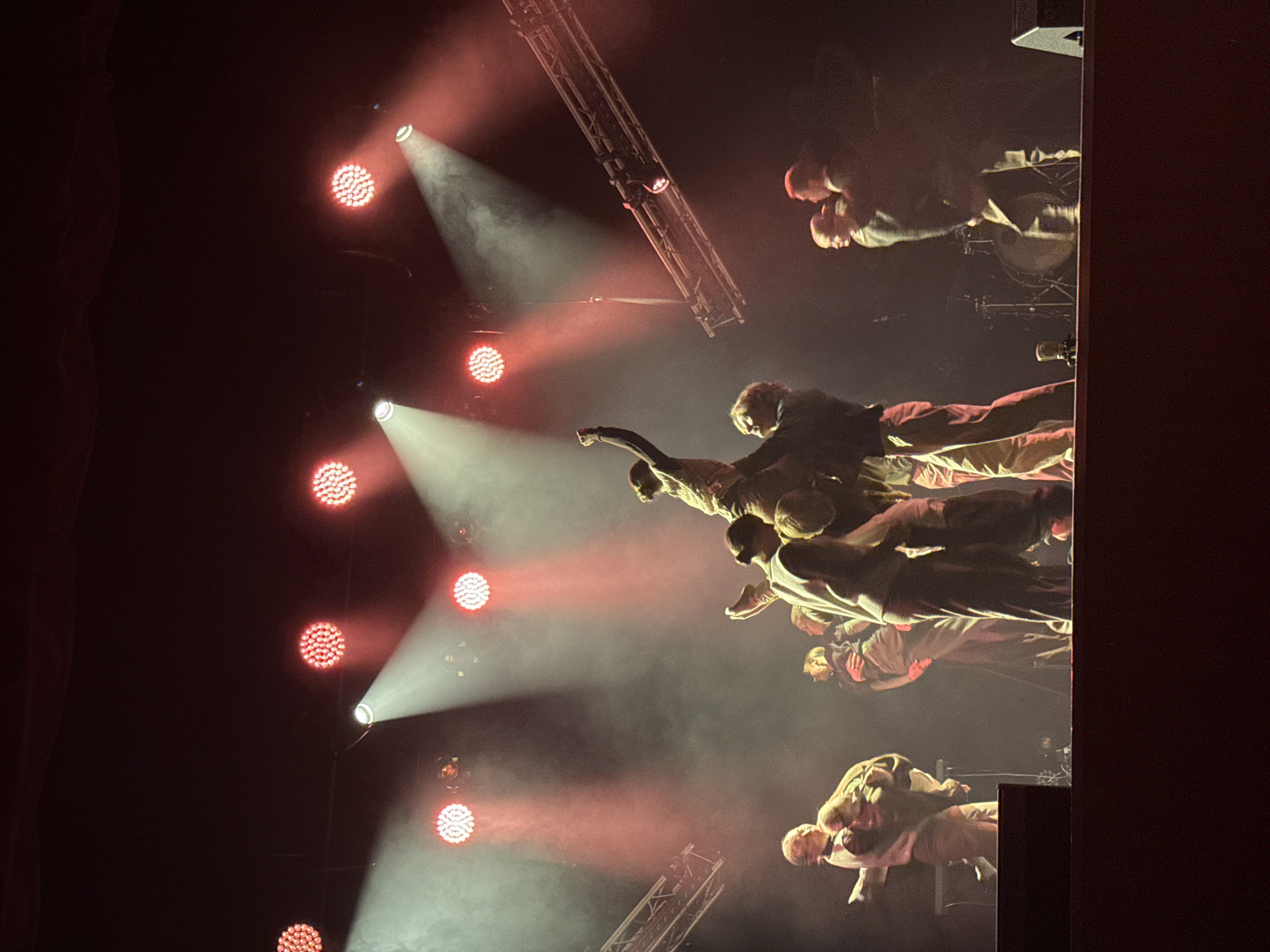 On a dimly lit stage in Perm, Russia, a group of performers are engaged in a dynamic dance routine. Red stage lights cast a warm glow, illuminating the fog that fills the air and accentuating the performers' movements. Several individuals are captured mid-air, lifted by their peers, showcasing strength and coordination. The performers are dressed in muted, contemporary clothing, suggesting a modern dance piece. In the background, stage lighting rigs and trusses are visible, along with a drum and microphone, hinting at a performance that may incorporate musical elements. The overall atmosphere is dramatic and artistic, with a focus on human connection and physical expression.