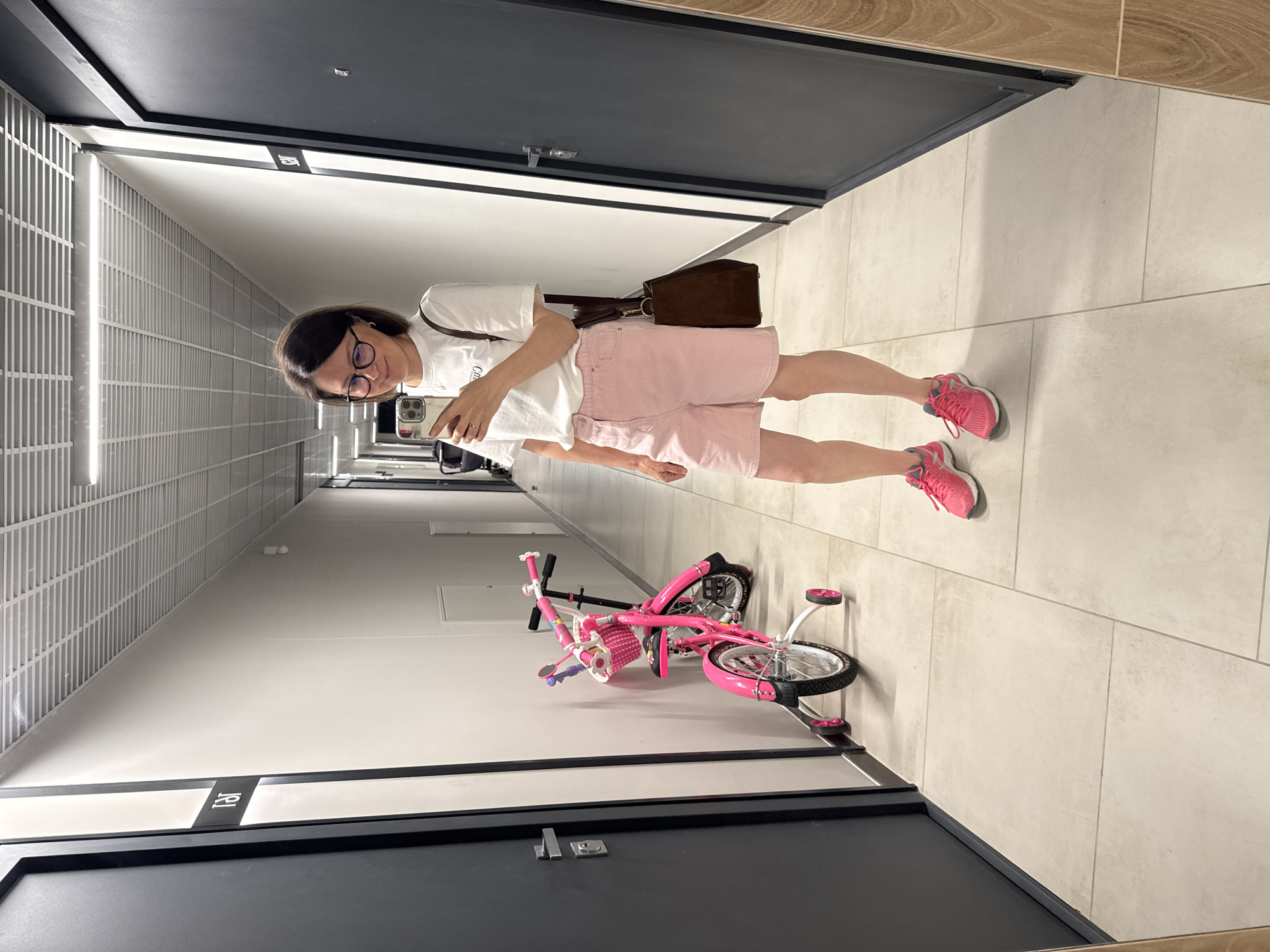 A woman stands in a brightly lit, modern hallway with cream-colored tiled floors and white walls. The hallway is lined with dark gray doors, each with a silver handle and a black and white sign above it. The woman, wearing glasses, a white t-shirt, and light pink shorts, is holding a smartphone in her right hand, taking a selfie. She has a brown bag slung over her left shoulder. To her left, a bright pink child's bicycle with training wheels is parked against the wall. The bicycle has a basket on the handlebars adorned with colorful decorations. The hallway appears to be in an apartment building or a similar residential complex. The overall aesthetic suggests a clean and organized environment. The location is specified as Izhevsk, Russia.
