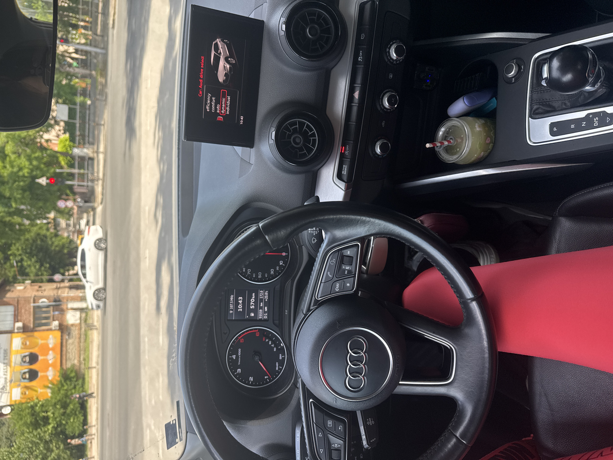 The image shows the interior of an Audi car, viewed from the driver's seat. The car is in motion, as indicated by the blurred motion of the exterior through the windshield. The dashboard displays the time as 10:43 and a digital odometer reading of 93559 km. The car's drive select system is set to "dynamic." The exterior scene visible through the windshield shows a street with trees and buildings, a white car driving in the opposite lane, and pedestrians on the sidewalk. The location is identified as Plovdiv, Bulgaria. The interior includes the steering wheel with controls, air vents, and the infotainment screen. A red-covered seat is visible in the foreground.