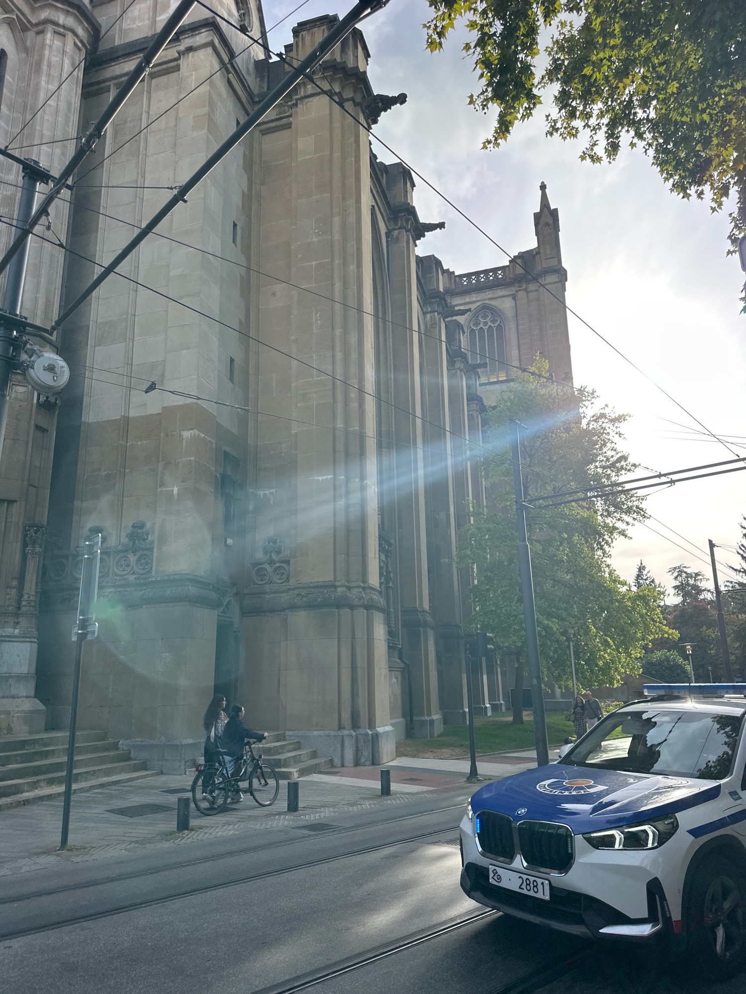 The image shows a police car in the city of Vitoria-Gasteiz, Spain. The police car, a white BMW with blue and white markings and a blue light bar, is positioned on a street with tram tracks. Behind the police car, a large stone building with architectural details resembling a cathedral or a historic public building is visible. The building has large windows and ornate carvings. In front of the building, two people are seen standing next to a bicycle on a paved sidewalk. Overhead, tram wires crisscross the sky, and the sun is casting lens flares across the scene. Trees with green foliage are also present, suggesting an urban park or streetscape. The ground surface includes tram tracks and a paved road. The overall impression is a street scene in a European city, with a focus on the police vehicle and the historic architecture.