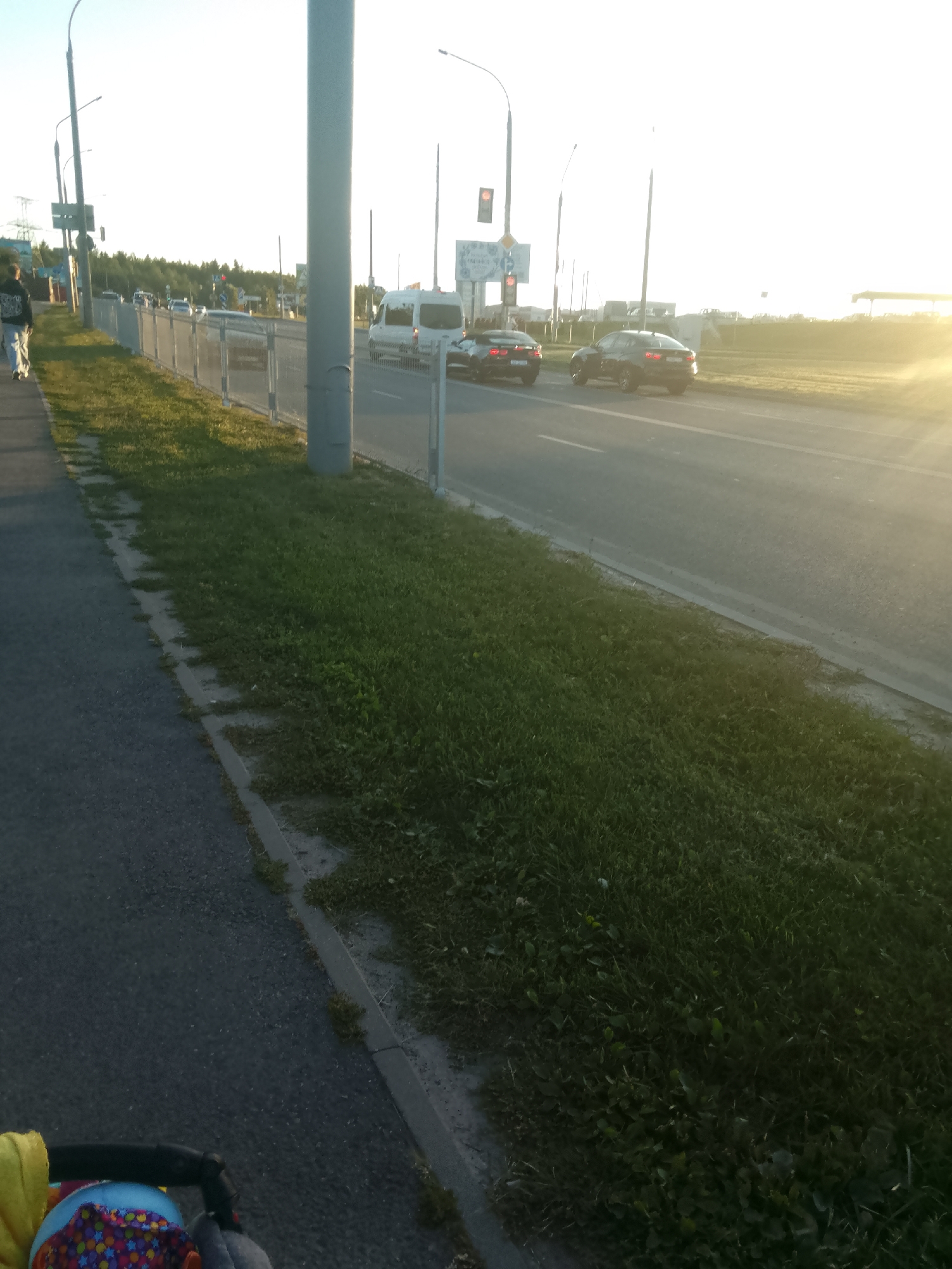 A daytime, eye-level, wide shot shows a paved walkway bordering a grassy area next to a road. In the foreground, a stroller is partially visible on the left. A person walks away from the camera on the walkway. A metal fence separates the walkway from the road, on which several cars are driving. A white van is also present on the road. Streetlights and traffic signals are visible in the background, along with trees and buildings. The sun is setting on the right side of the frame, casting a warm glow. The scene appears to be located in the Shchomyslitsky Selsoviet, Belarus.