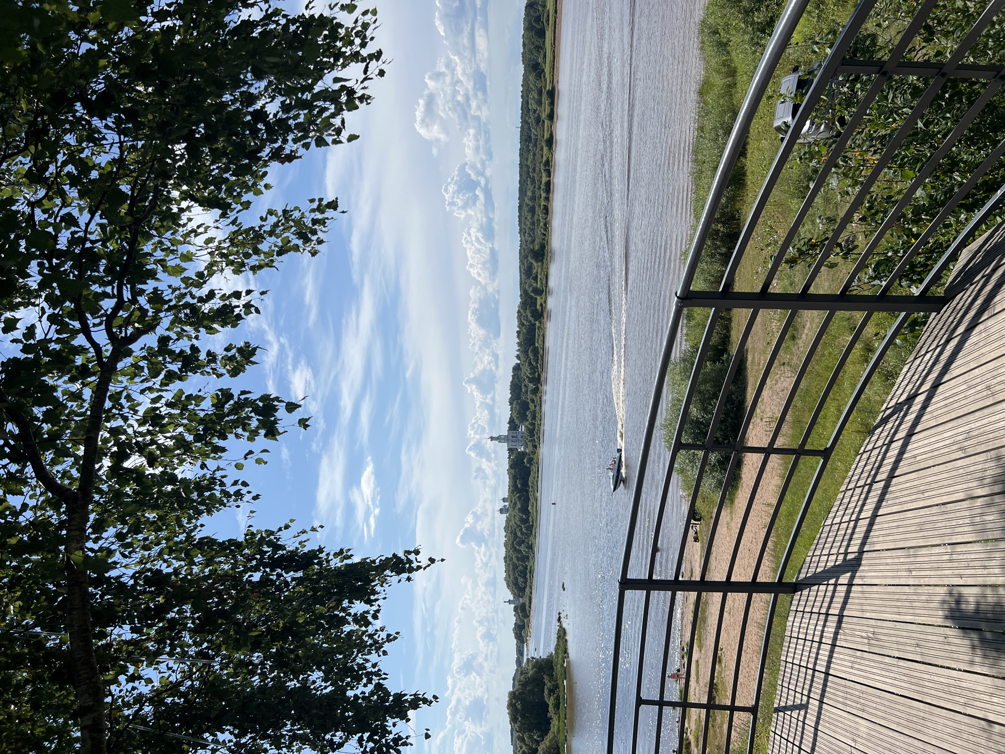 An exterior daytime shot overlooks a wide, calm river in Veliky Novgorod, Russia. The foreground features a wooden deck with a metal railing, casting shadows from unseen foliage above. To the left of the railing, a sandy beach is visible with a few individuals. On the river, a small motorboat travels at speed, leaving a wake. The opposite bank of the river is lined with dense green trees. In the distance, across the river, several white domed structures, likely churches, are visible against a bright blue sky dotted with white, fluffy clouds. The overall scene depicts a serene riverside landscape on a clear day.